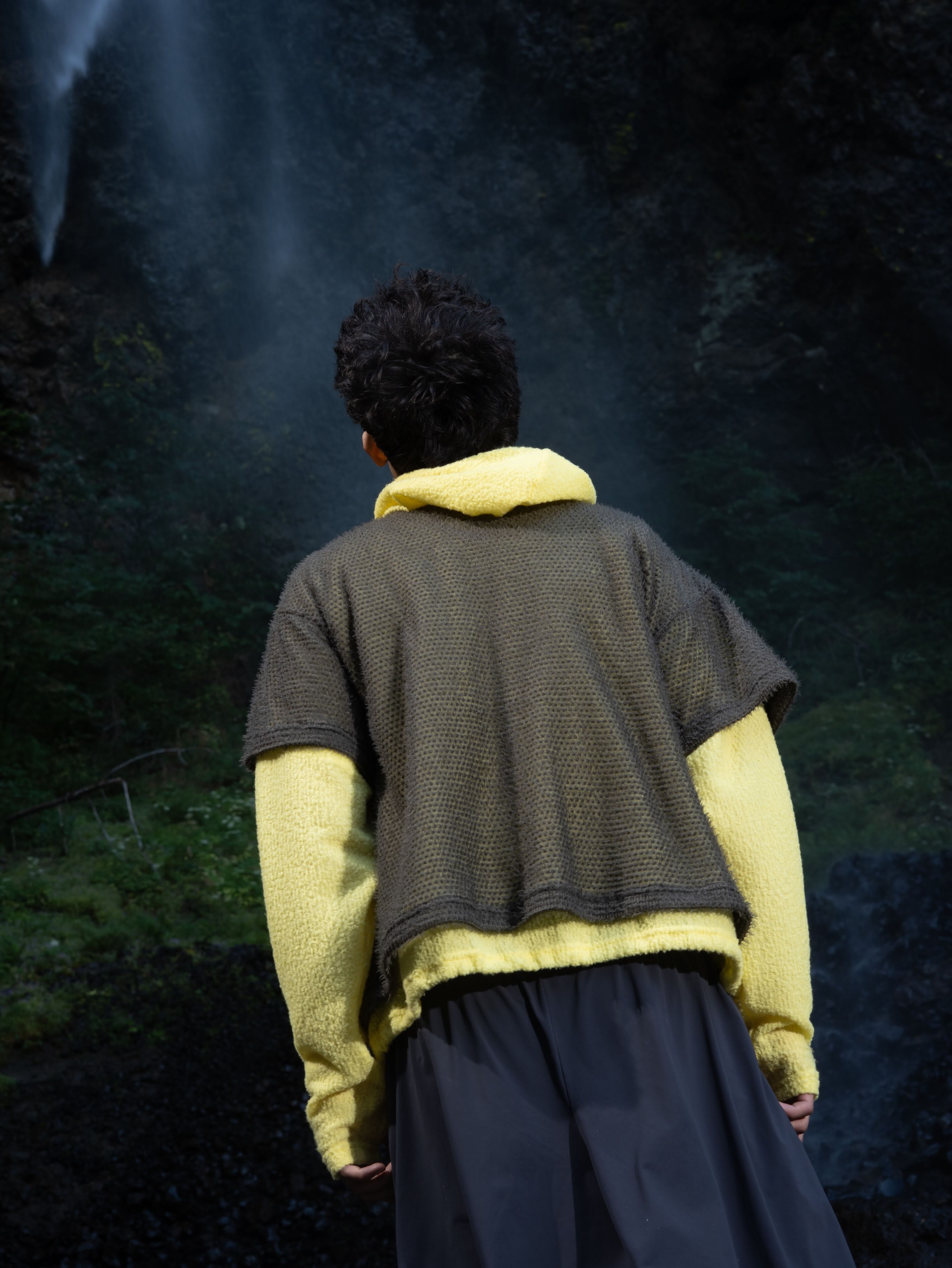 A man in a yellow sweater and olive-green textured overlay stands with his back to the camera against a dark, rocky background with a waterfall.
