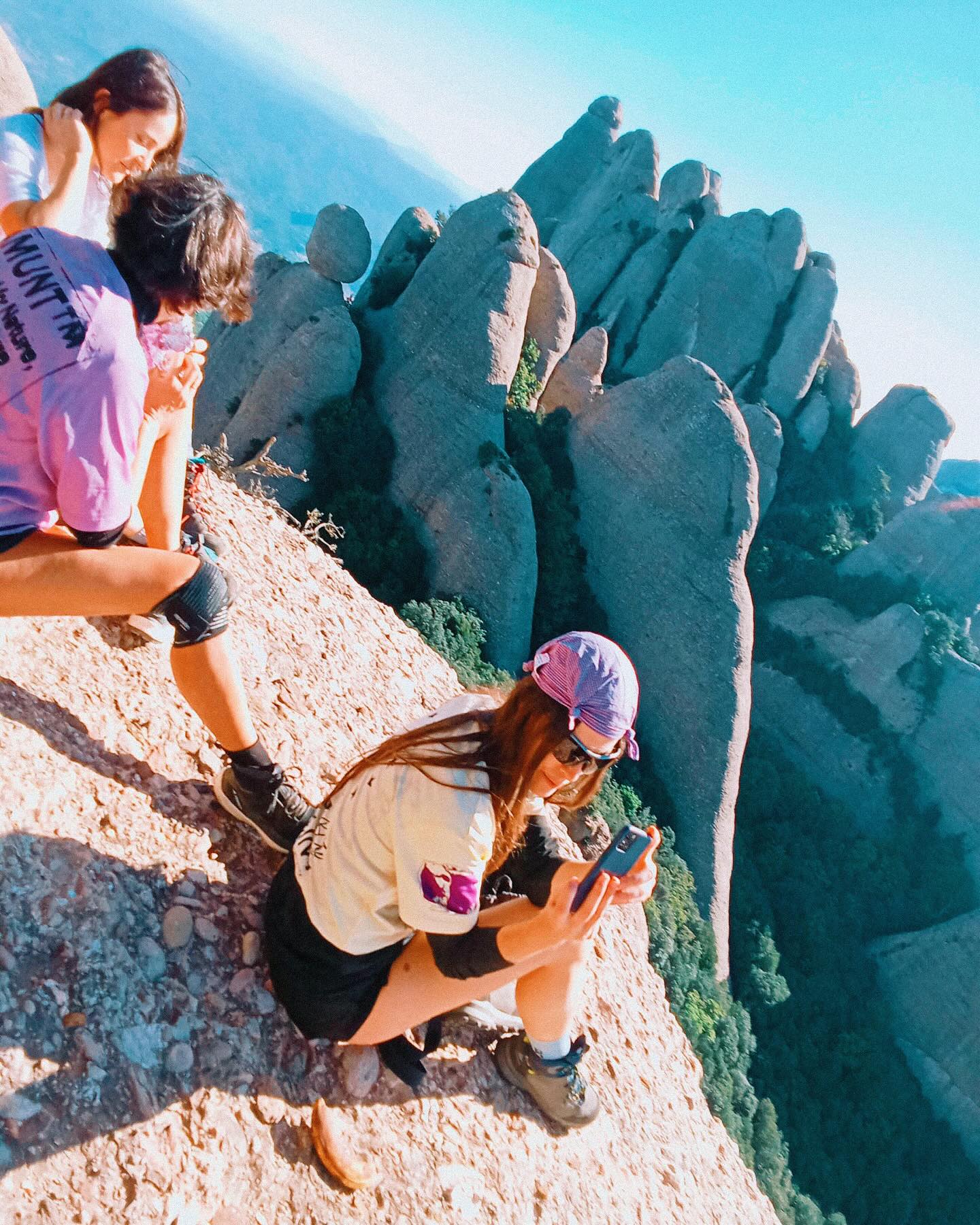 Two people sit on a steep, rocky cliffside in the foreground, while jagged mountain peaks rise against a clear blue sky in the background.