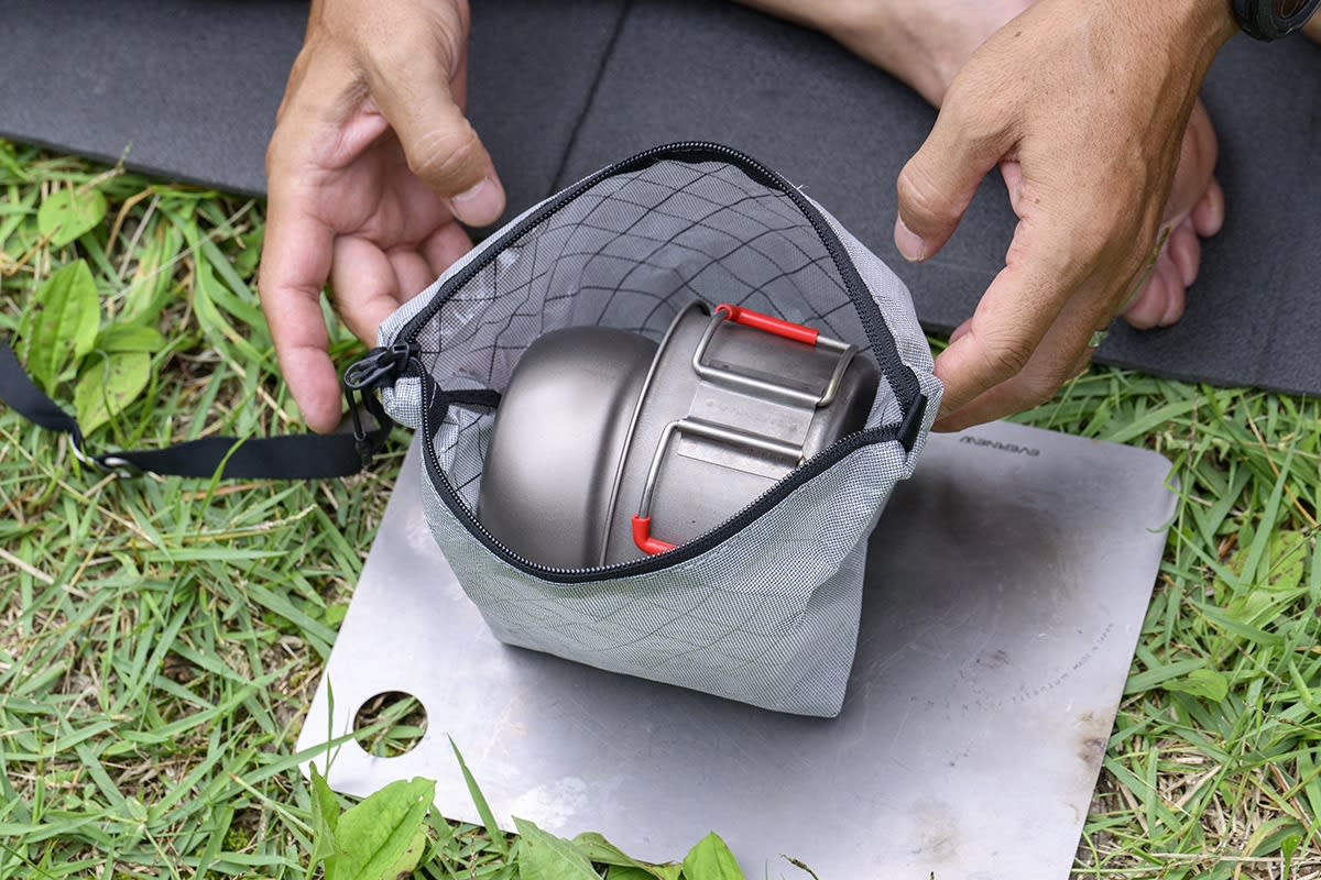 A person's hands hold open a grey mesh pouch containing a metal camping pot, resting on a silver tray over green grass and a black mat.