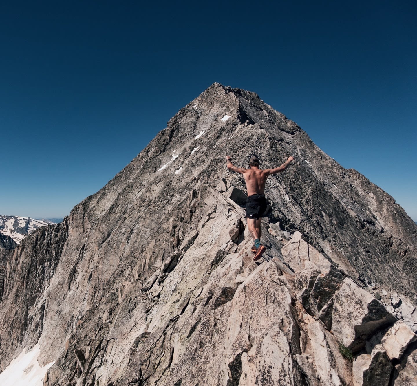 A shirtless man stands with arms raised on a jagged rocky ridge, facing a towering mountain peak under a clear blue sky.