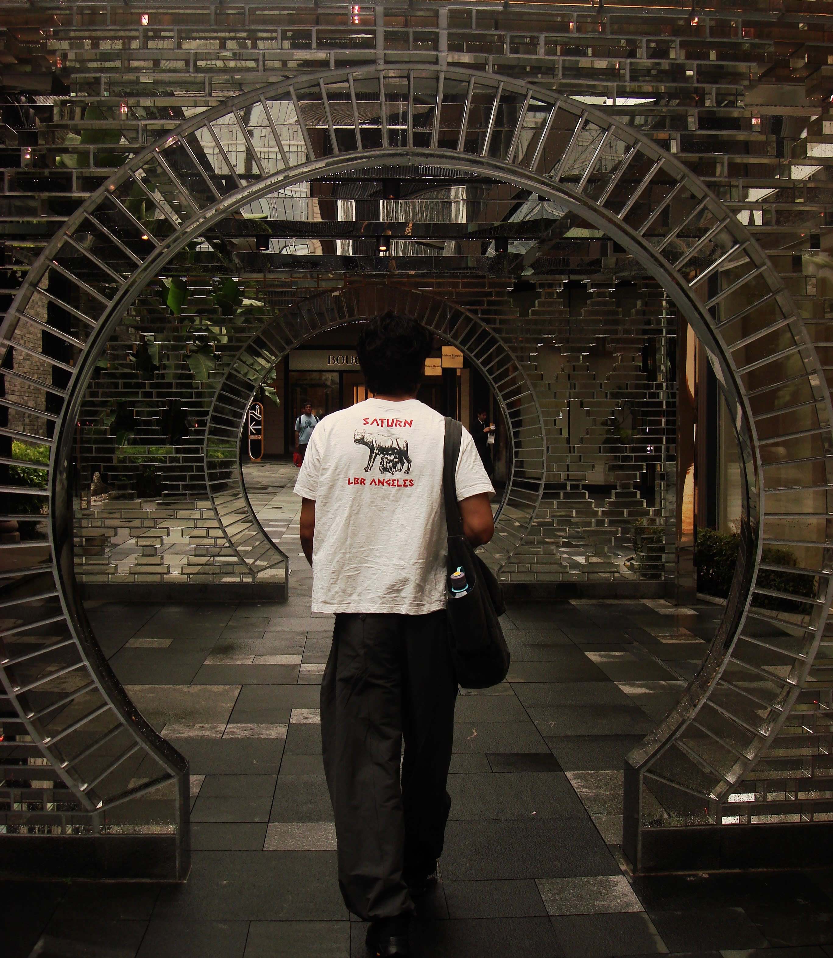 A man in a white t-shirt and dark pants walks through a series of large, metallic circular arches in a modern architectural space.