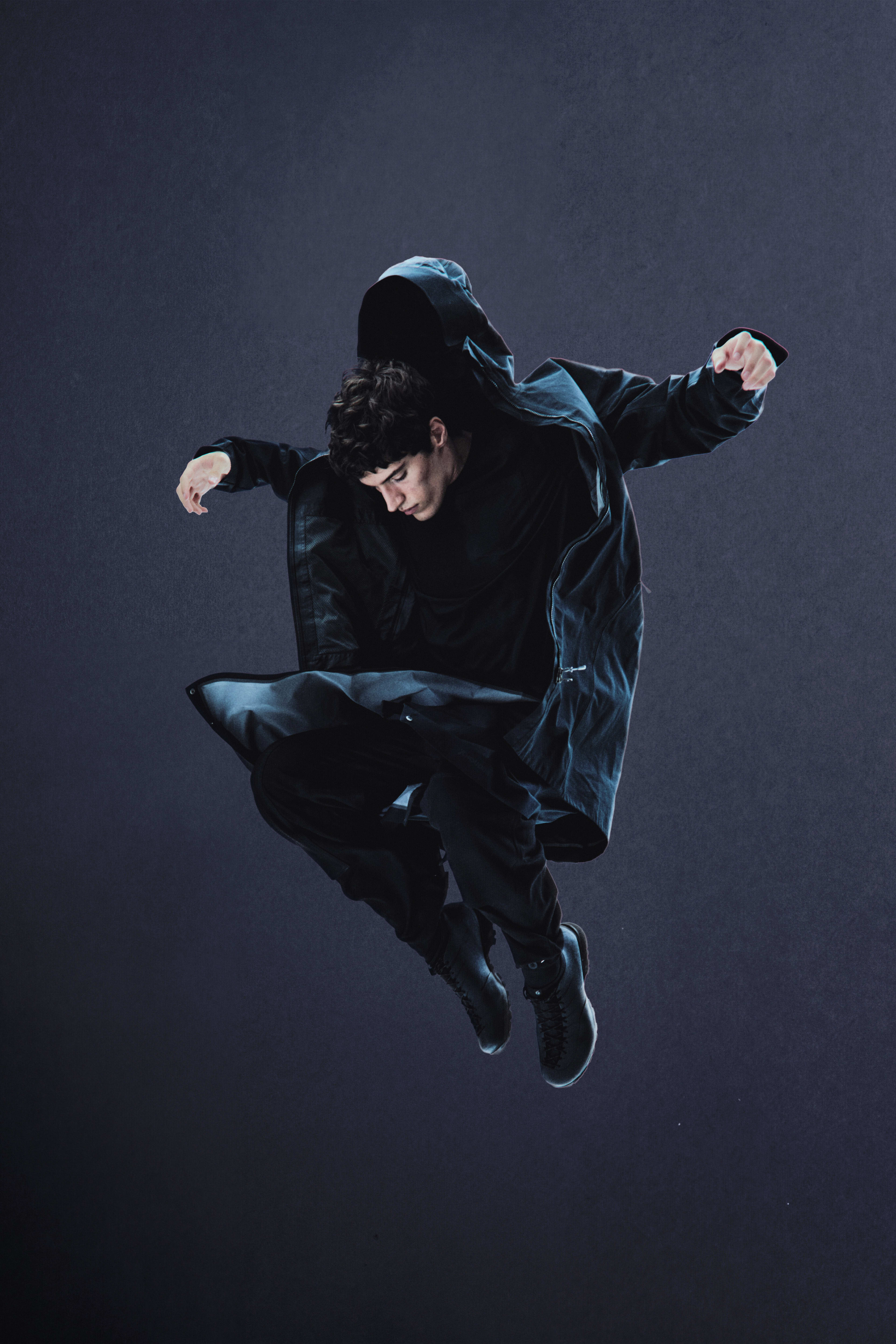 A man in a black hooded jacket and pants jumps mid-air against a dark, moody grey background.