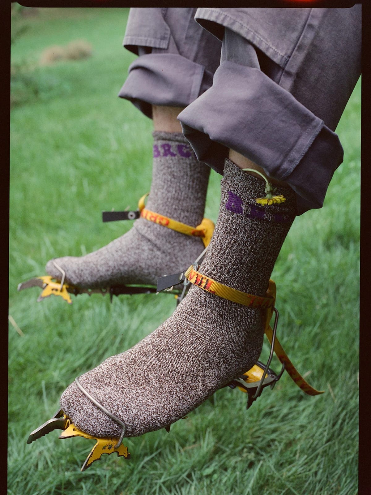 A person's feet, clad in thick socks and attached to crampons, rest on a vibrant green grassy field.