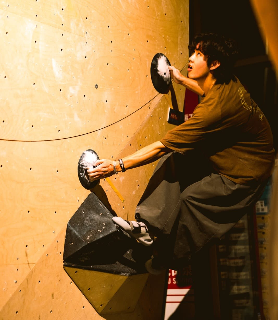 A man in a brown shirt climbs a wooden bouldering wall, gripping black holds against a dark, dimly lit background.