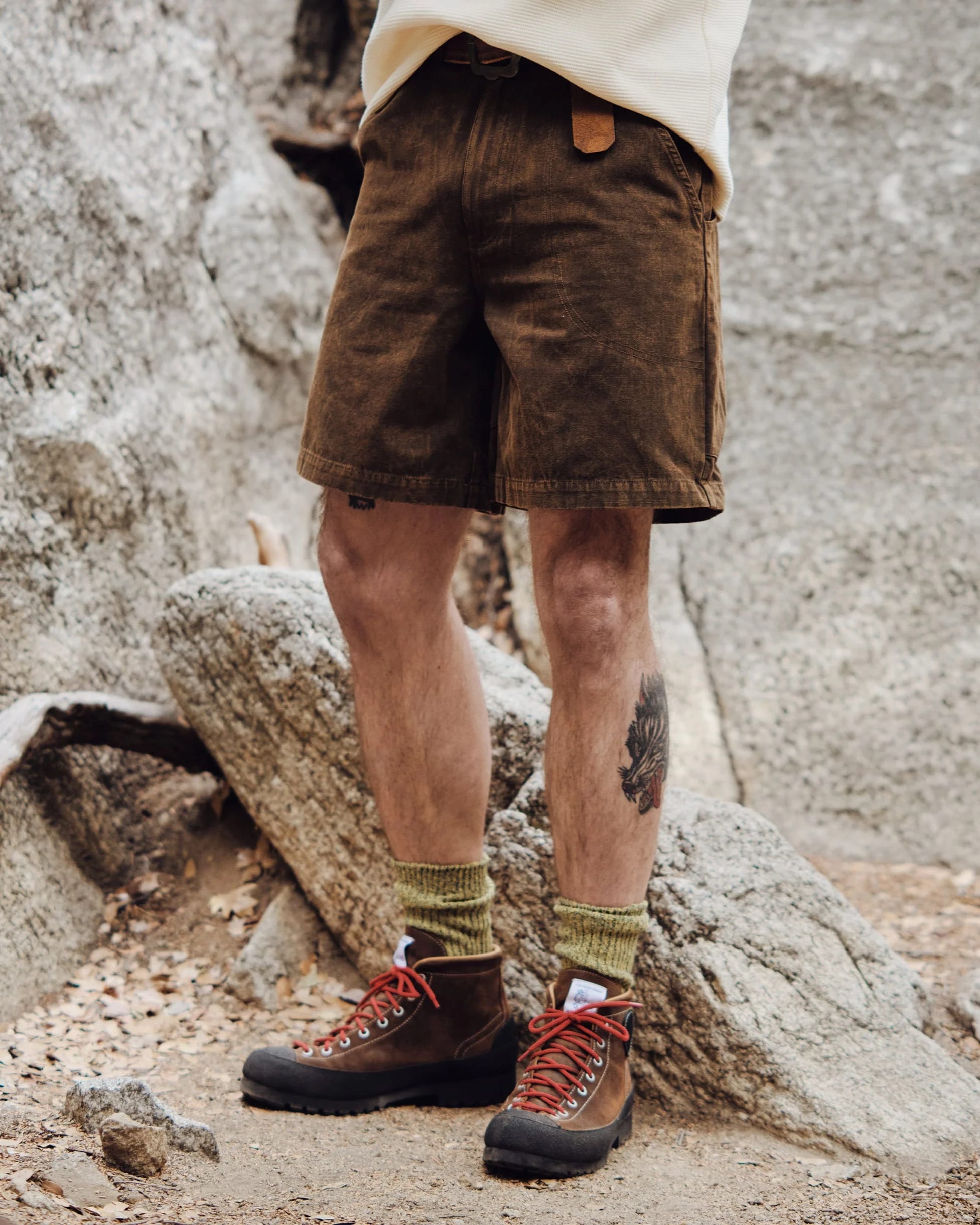 A person wearing brown corduroy shorts, green socks, and hiking boots stands on a rocky path with large boulders in the background.
