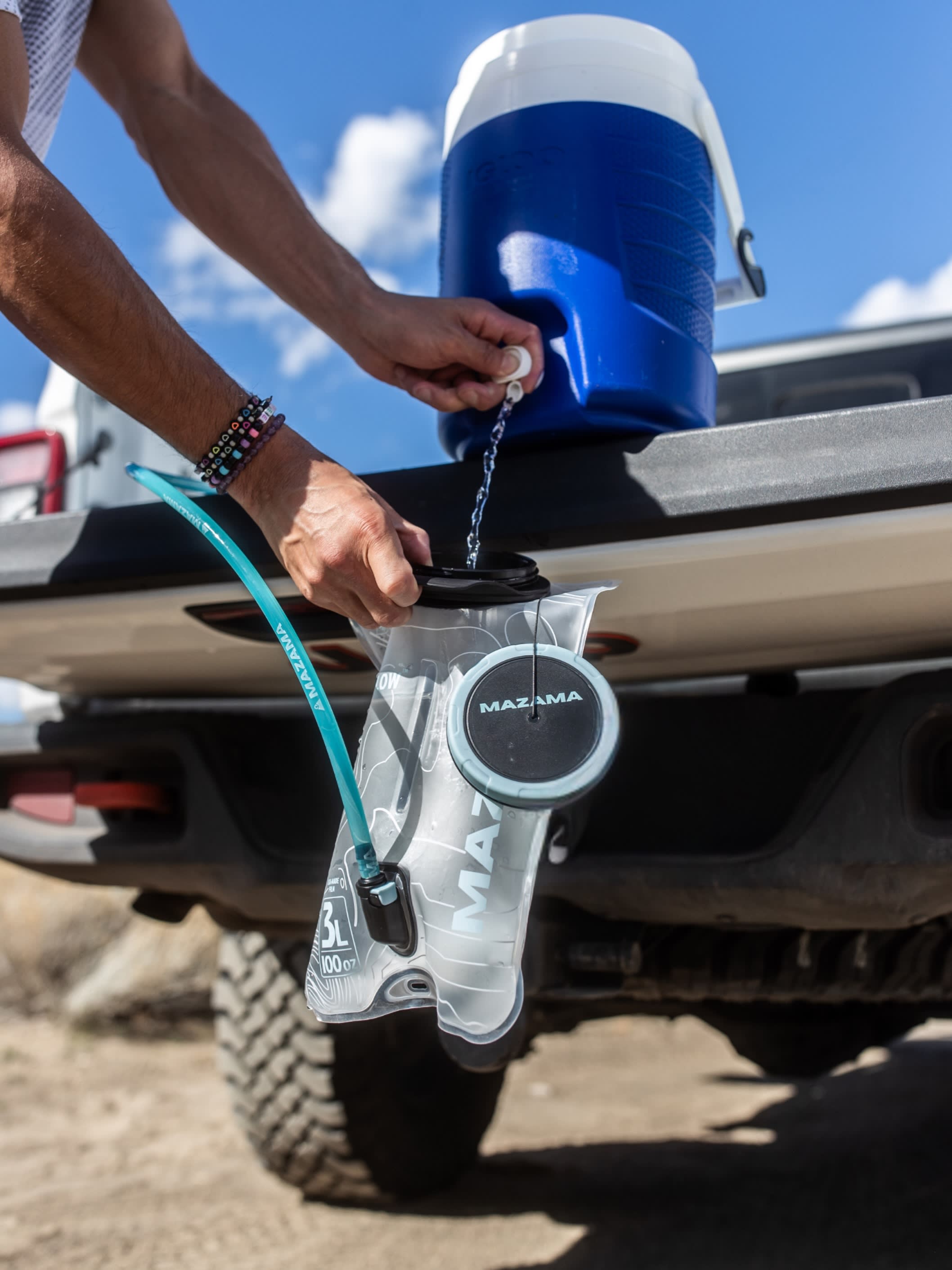 A person fills a clear hydration bladder from a blue water jug resting on the tailgate of a truck under a bright blue sky.