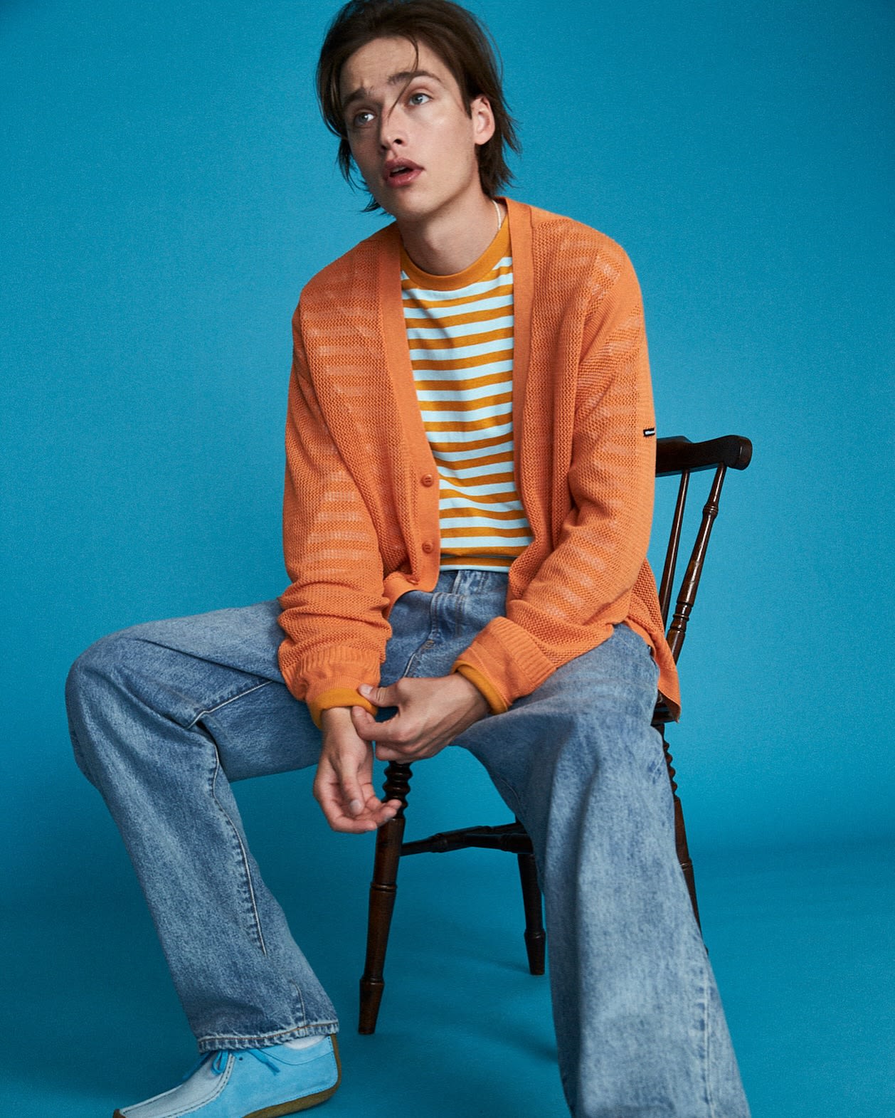 A young man in a striped shirt and orange cardigan sits on a chair against a bright blue background.