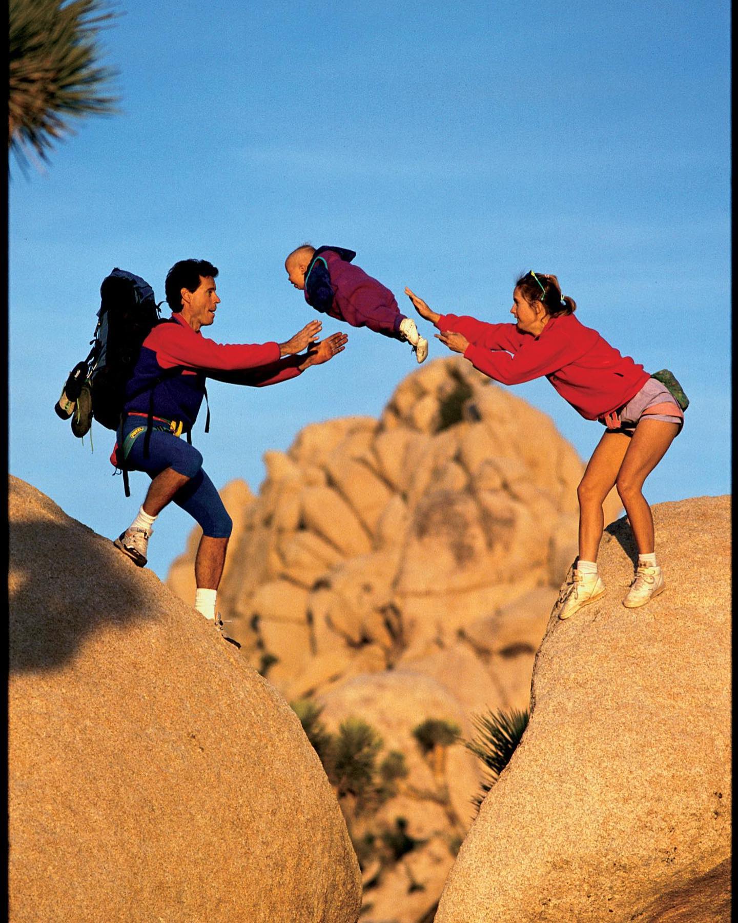 A man and woman toss a baby between them on a rocky outcrop with large boulders and a clear blue sky in the background.