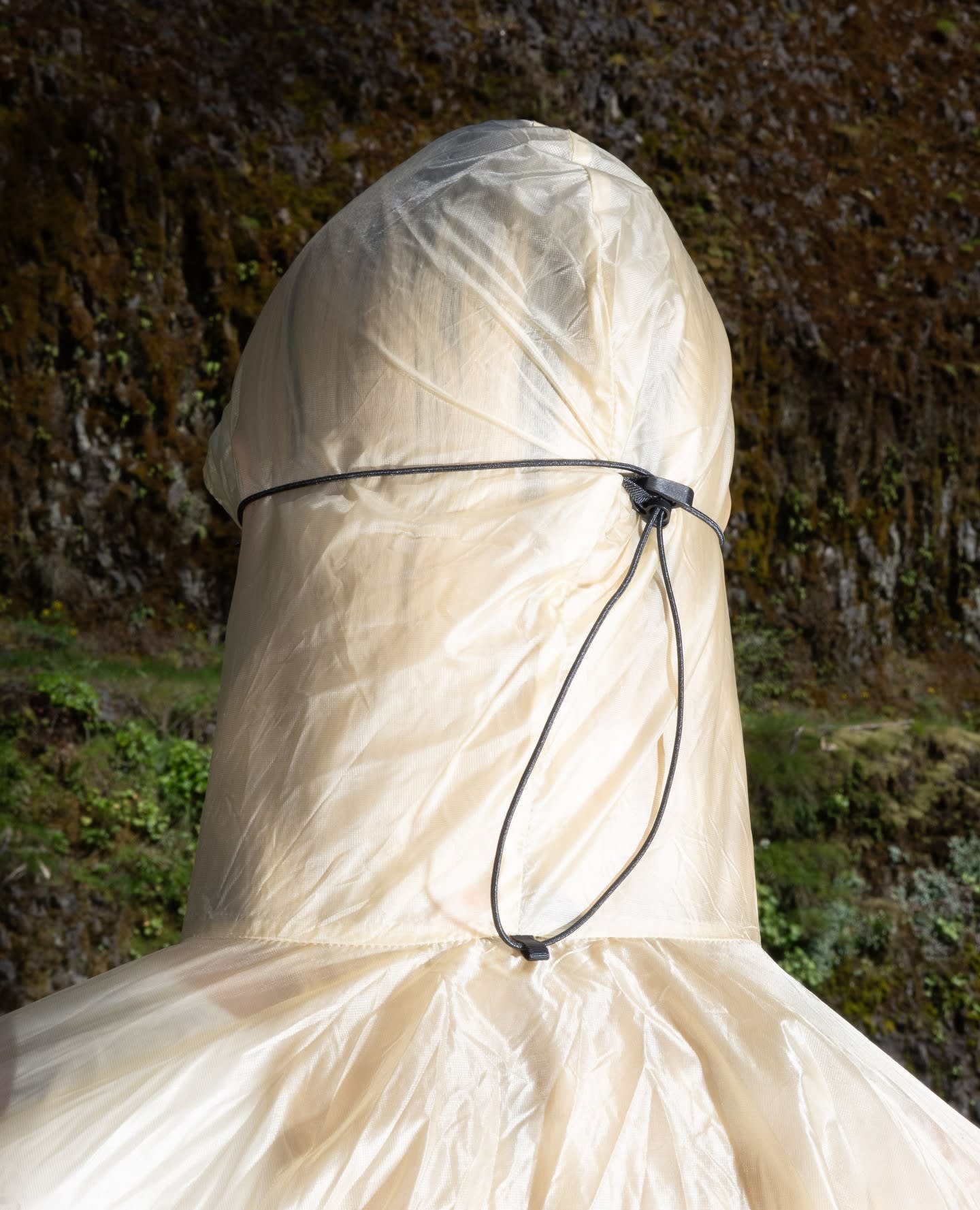 A person wearing a translucent white protective hood and gown stands before a dark, mossy rock wall.