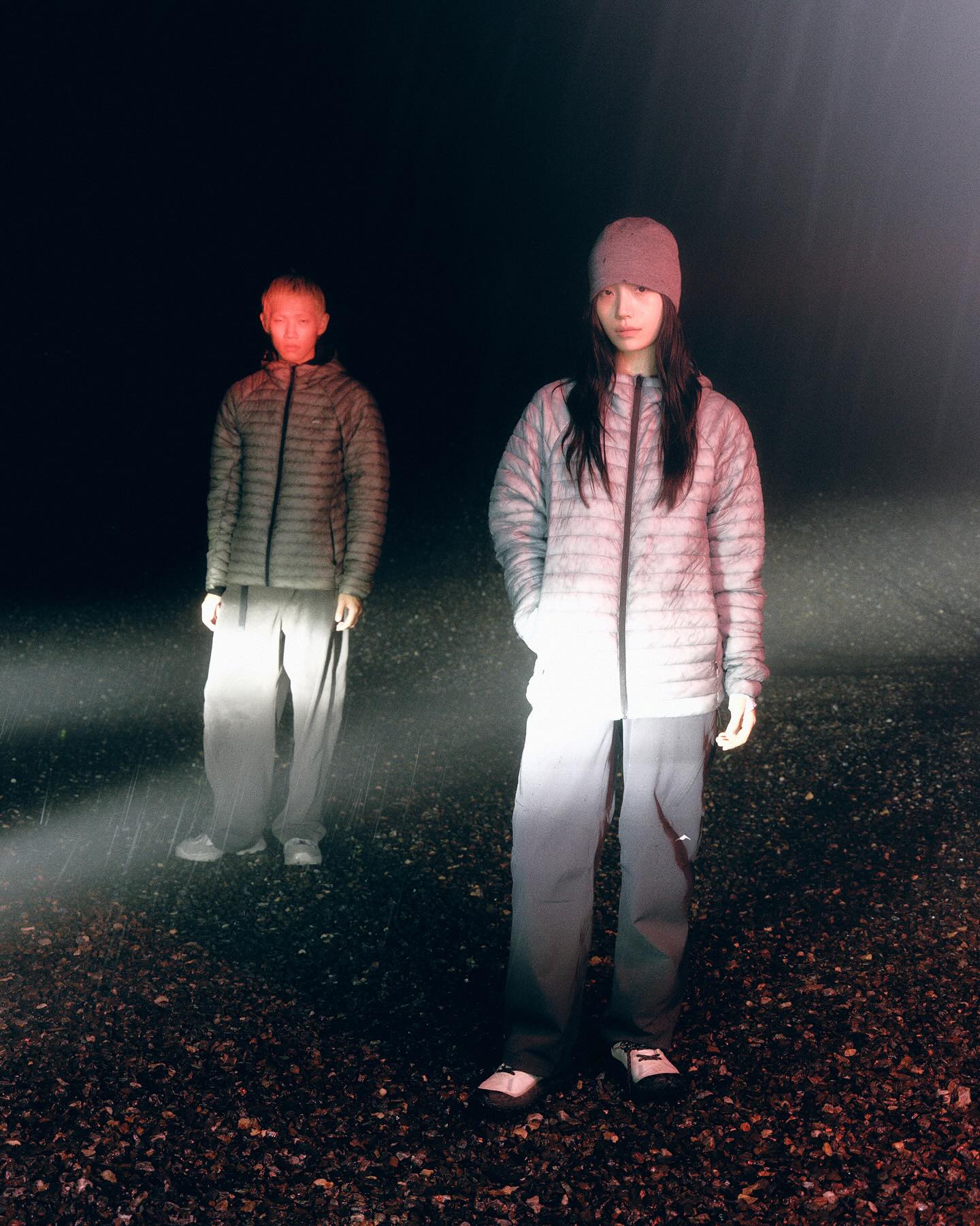 Two people wearing puffer jackets and grey pants stand on a dark, pebbled ground under a moody, dimly lit sky.