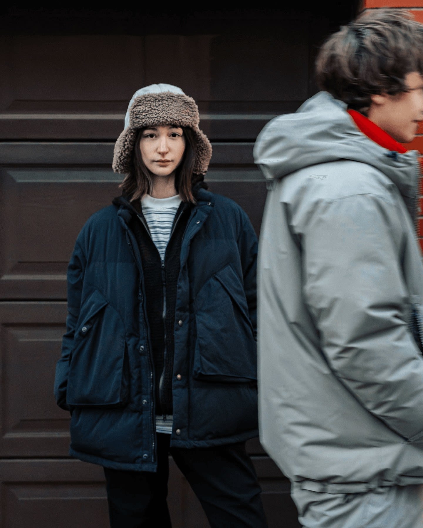 A young woman in a fuzzy hat and dark puffer jacket stands in front of a brown garage door, while a person in a light gray jacket walks past in the blurred background.
