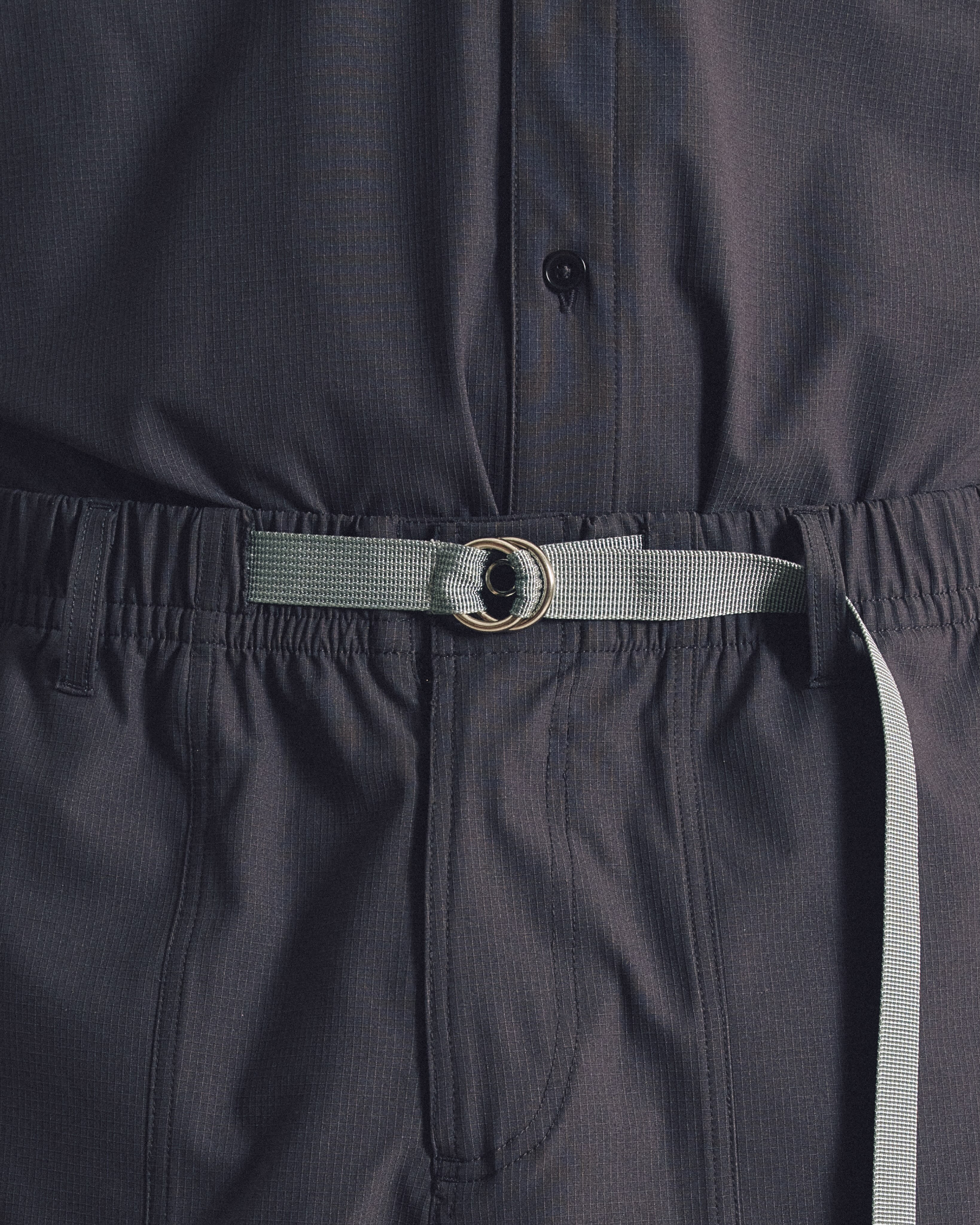 A grey fabric belt with a metal ring cinches the waist of dark grey textured trousers and a matching shirt.