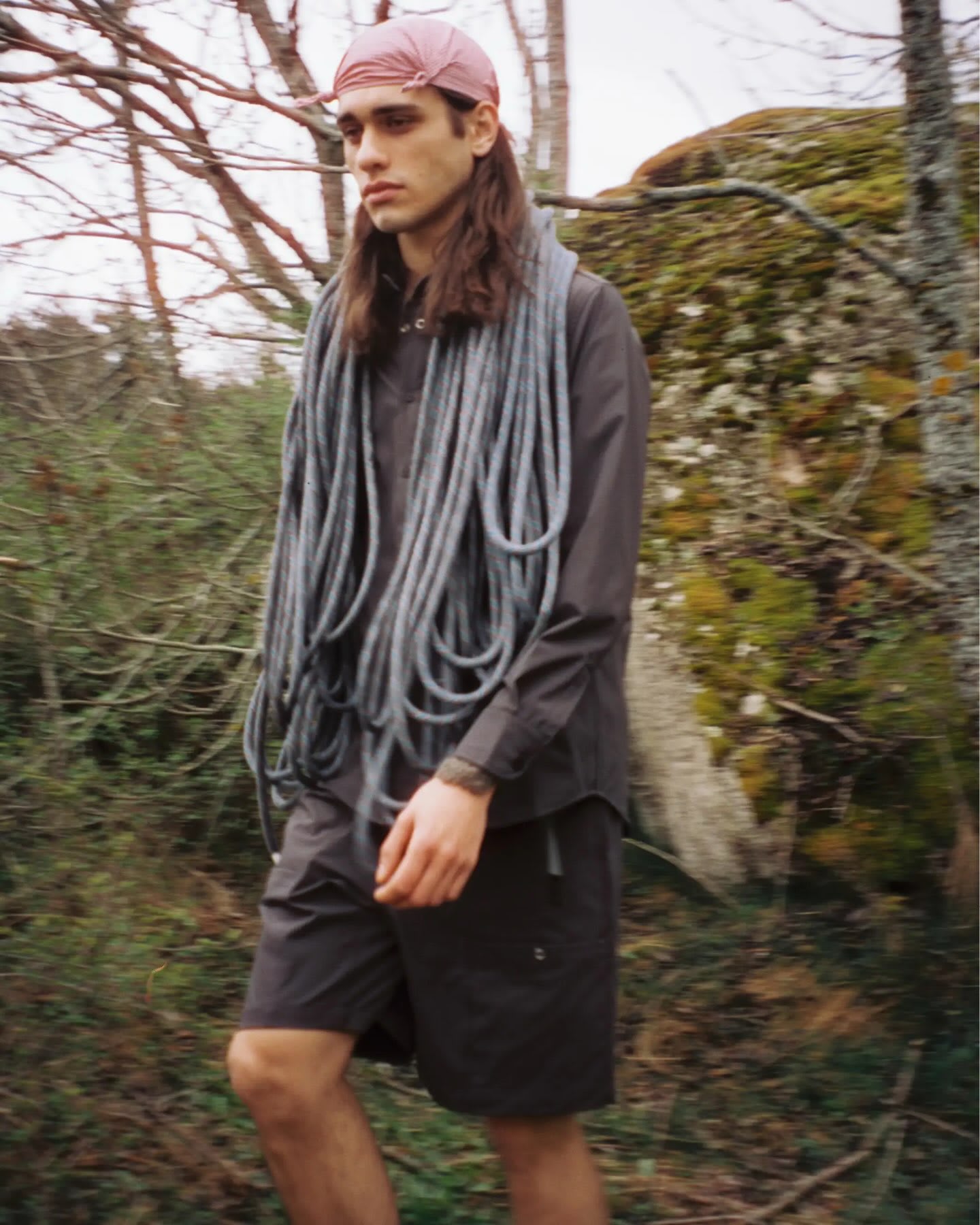 A man wearing a pink cap and a thick rope scarf stands in a wooded area with mossy rocks and bare branches.