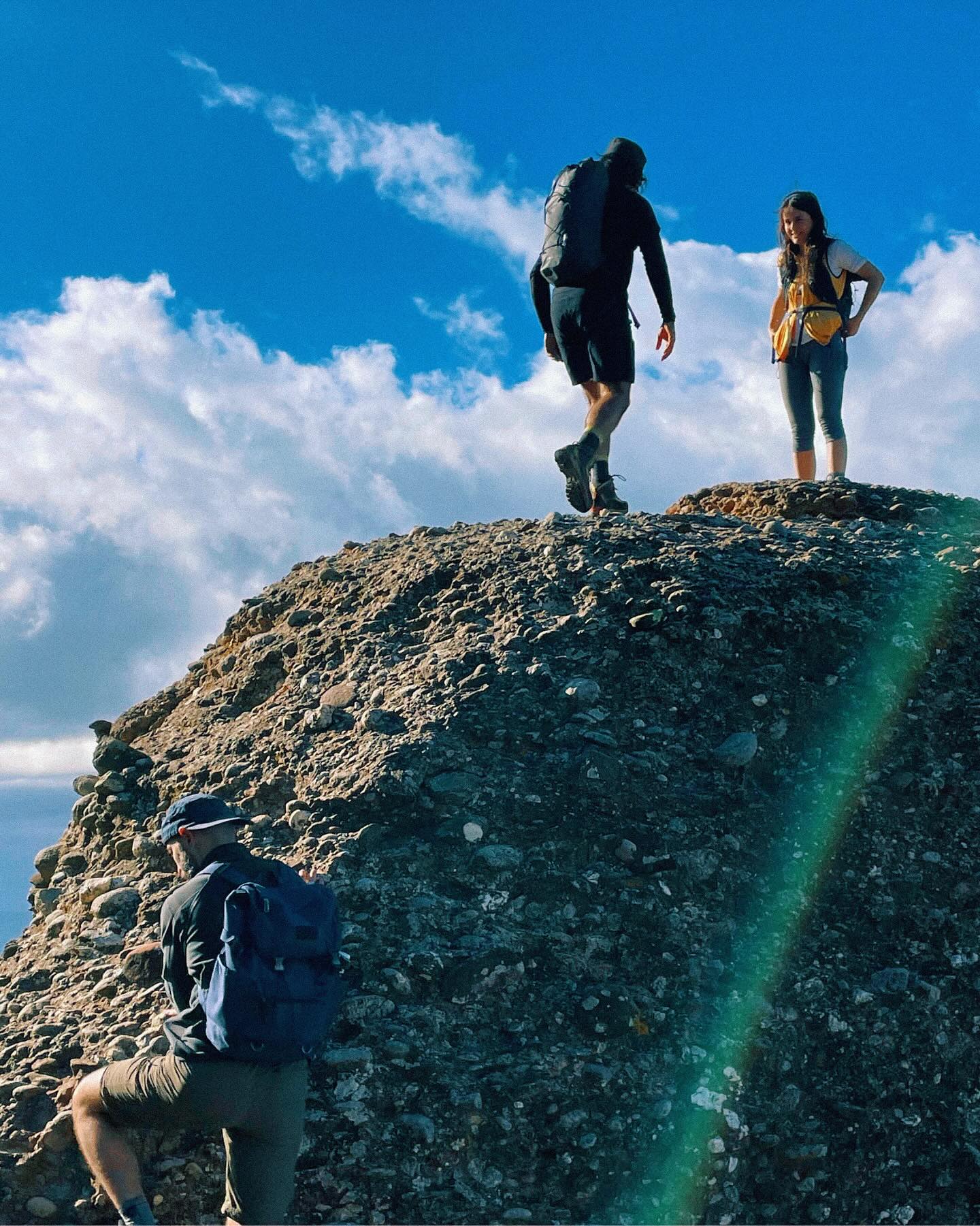 Three hikers with backpacks stand and climb a rocky peak under a bright blue sky with white clouds.
