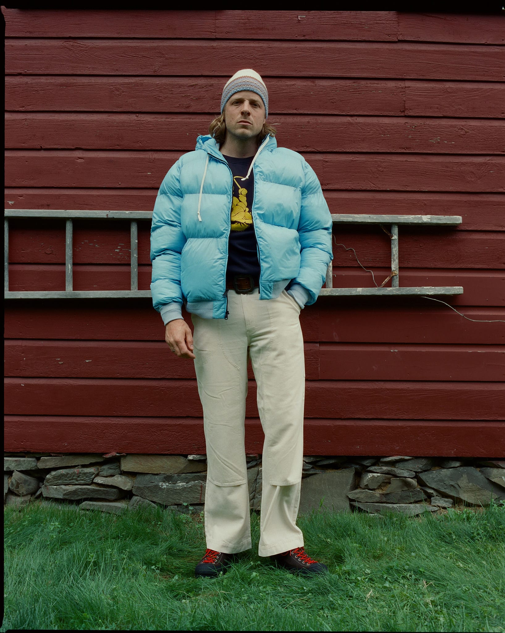 A man in a light blue puffer jacket and cream pants stands in front of a red wooden wall with a ladder, on a grassy area.