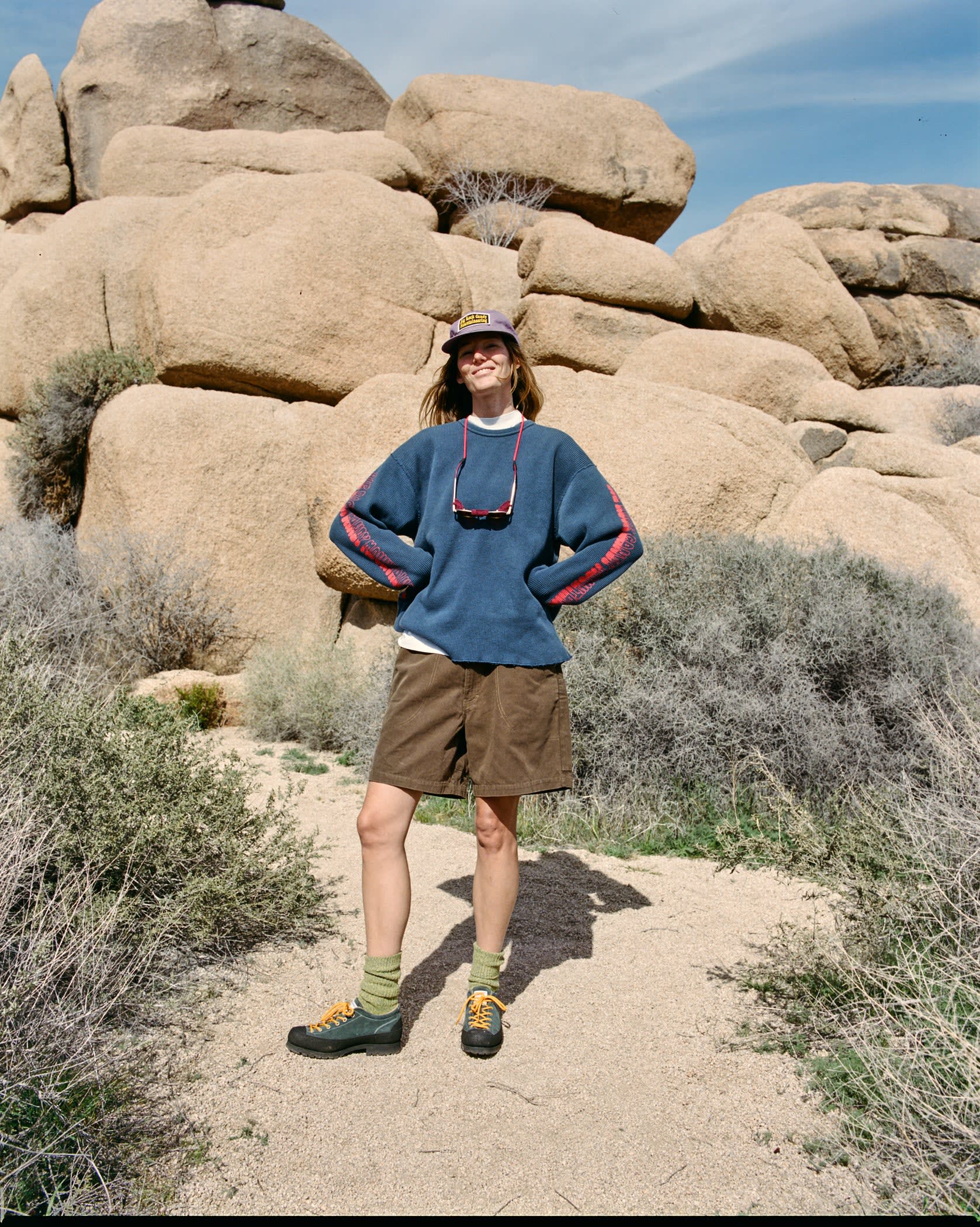 A person stands on a sandy path in the foreground, with large, rounded rock formations and sparse desert vegetation in the background.