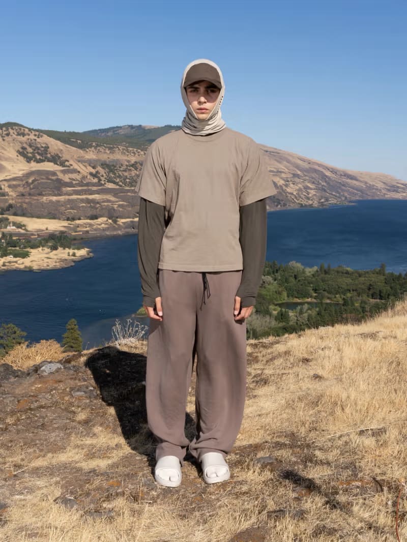 A man wearing a graphic white t-shirt, grey long-sleeved undershirt, and dark trousers stands on a sandy beach against a backdrop of dark, rocky cliffs.