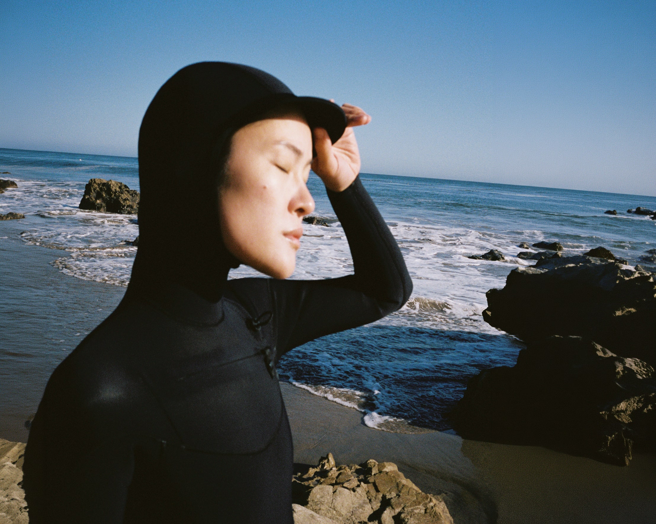 A person in a black wetsuit with a hood stands in the foreground against a background of a rocky coastline and blue ocean.