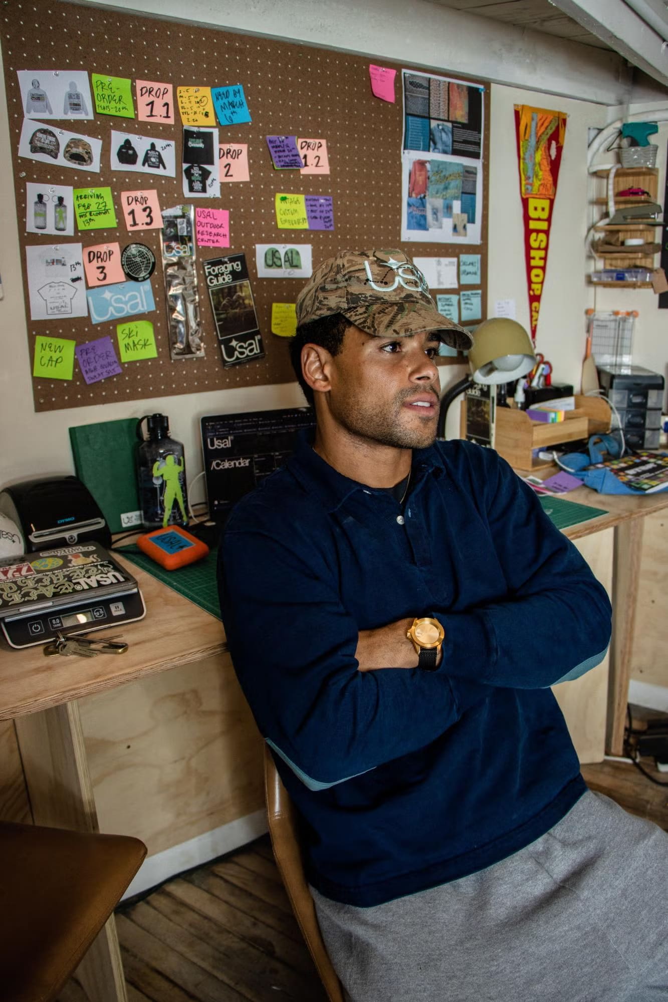 A man wearing a camouflage hat and navy blue shirt sits with crossed arms in front of a corkboard filled with notes and sketches.