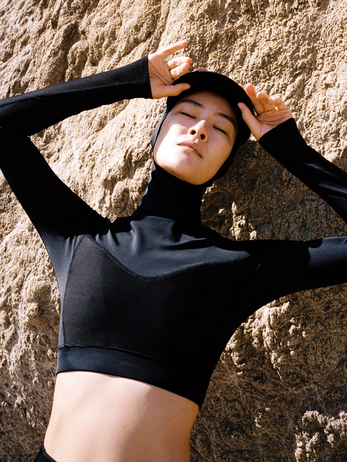 A person wearing a black long-sleeved crop top and headgear leans against a textured, rocky cliffside.