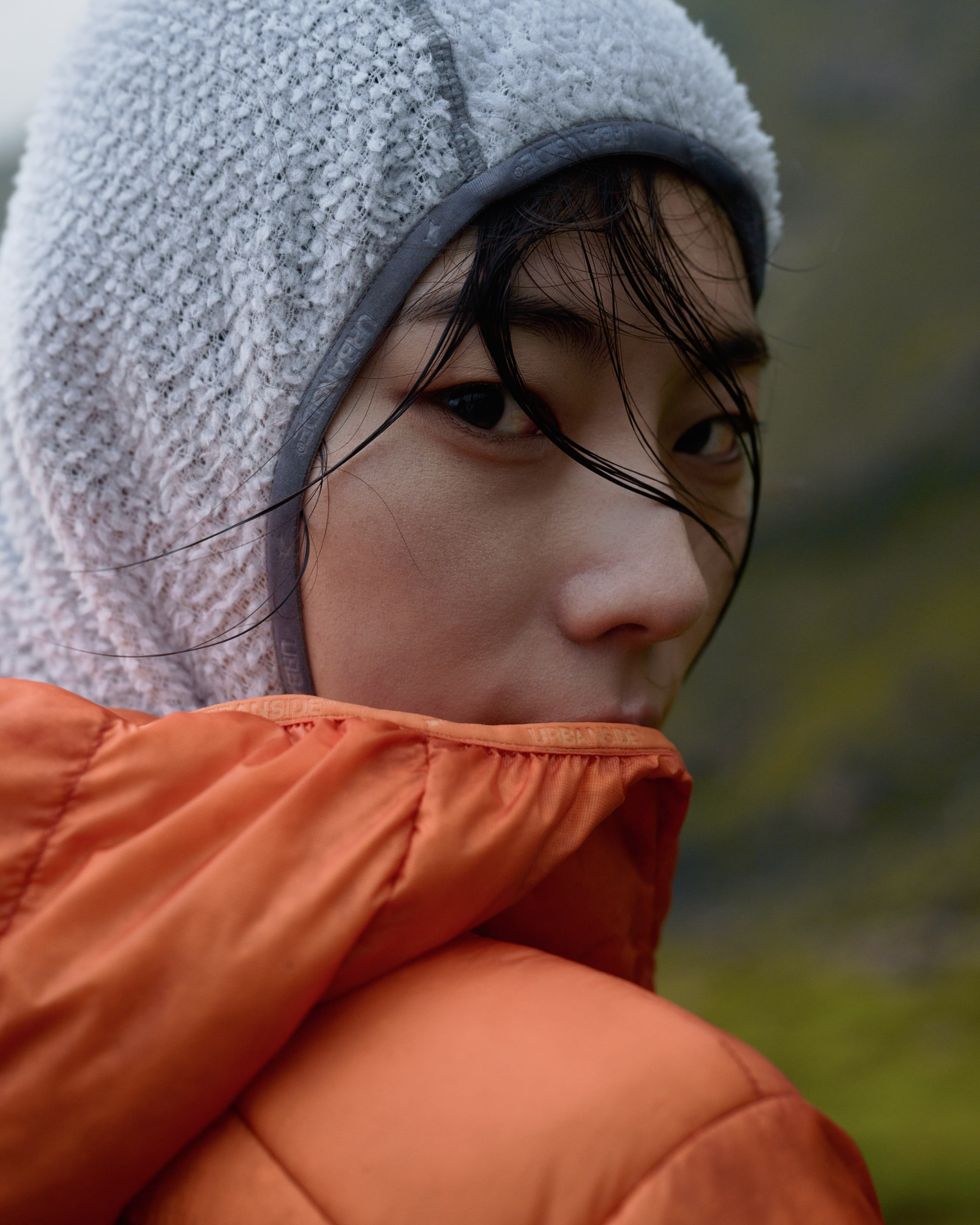 A person wearing a white textured beanie and an orange puffer jacket looks forward against a blurred green background.