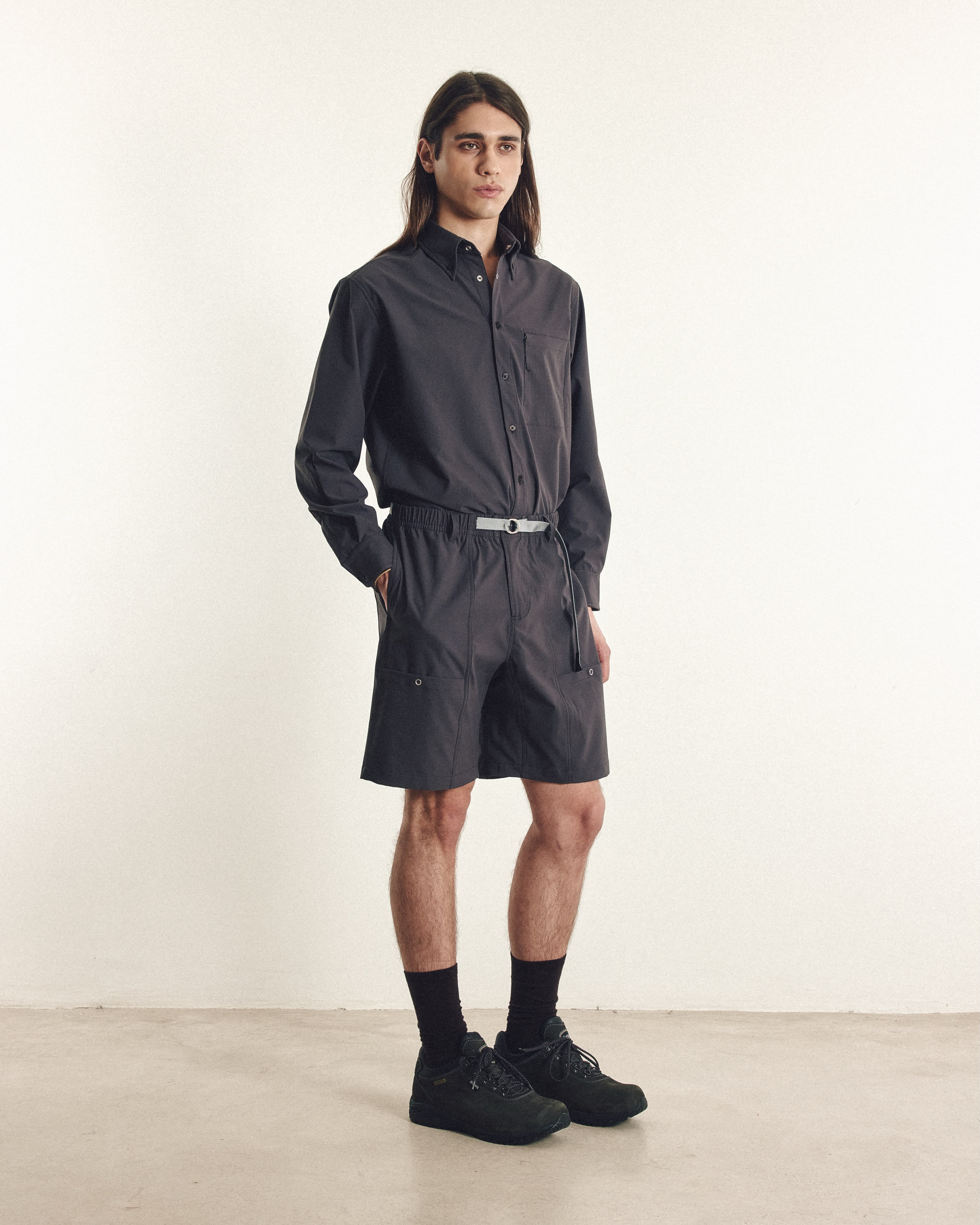 A man wearing a dark grey button-down shirt and matching shorts stands against a plain white wall.