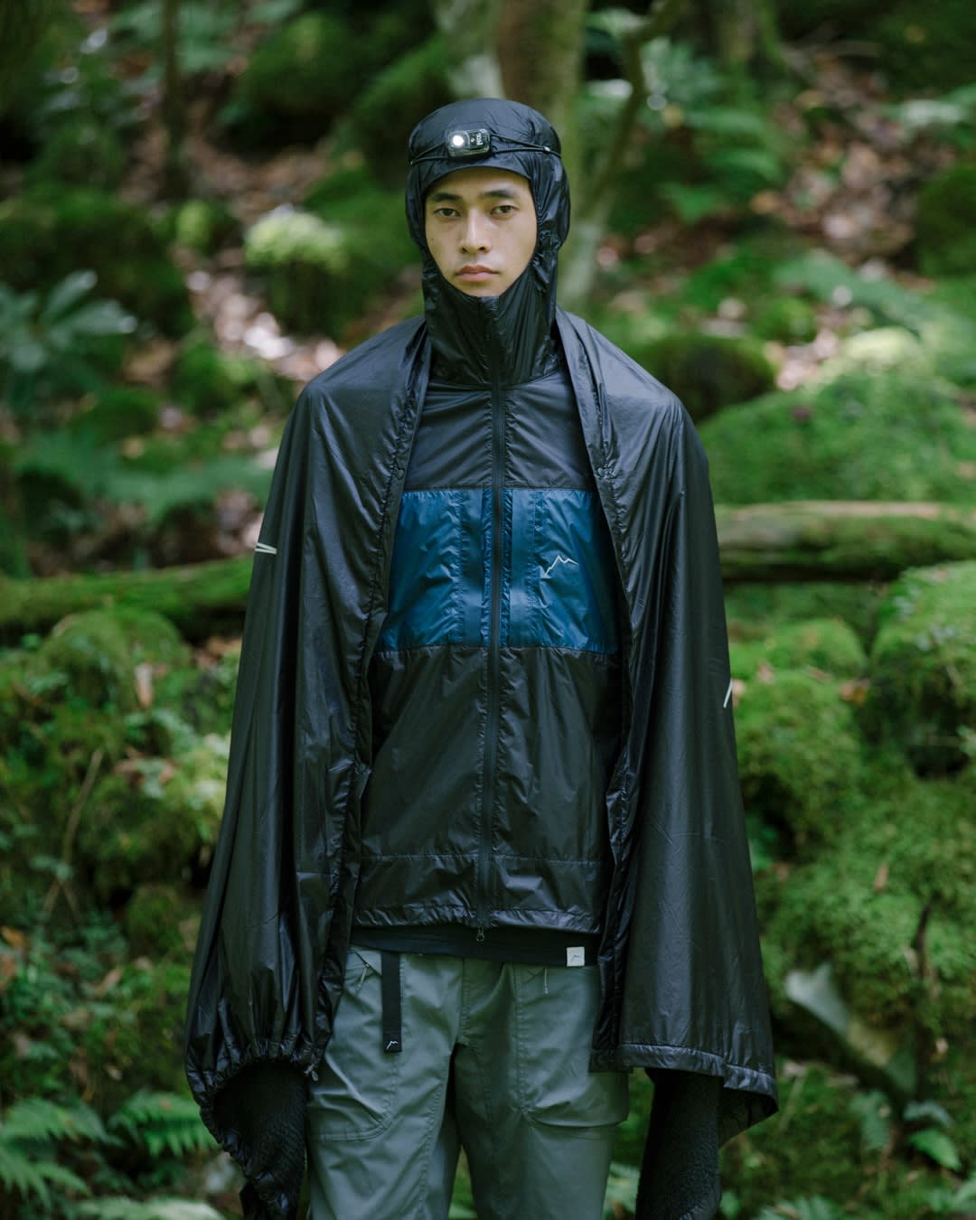 A man wearing a black and blue technical outdoor outfit and a headlamp stands in a lush, mossy green forest.