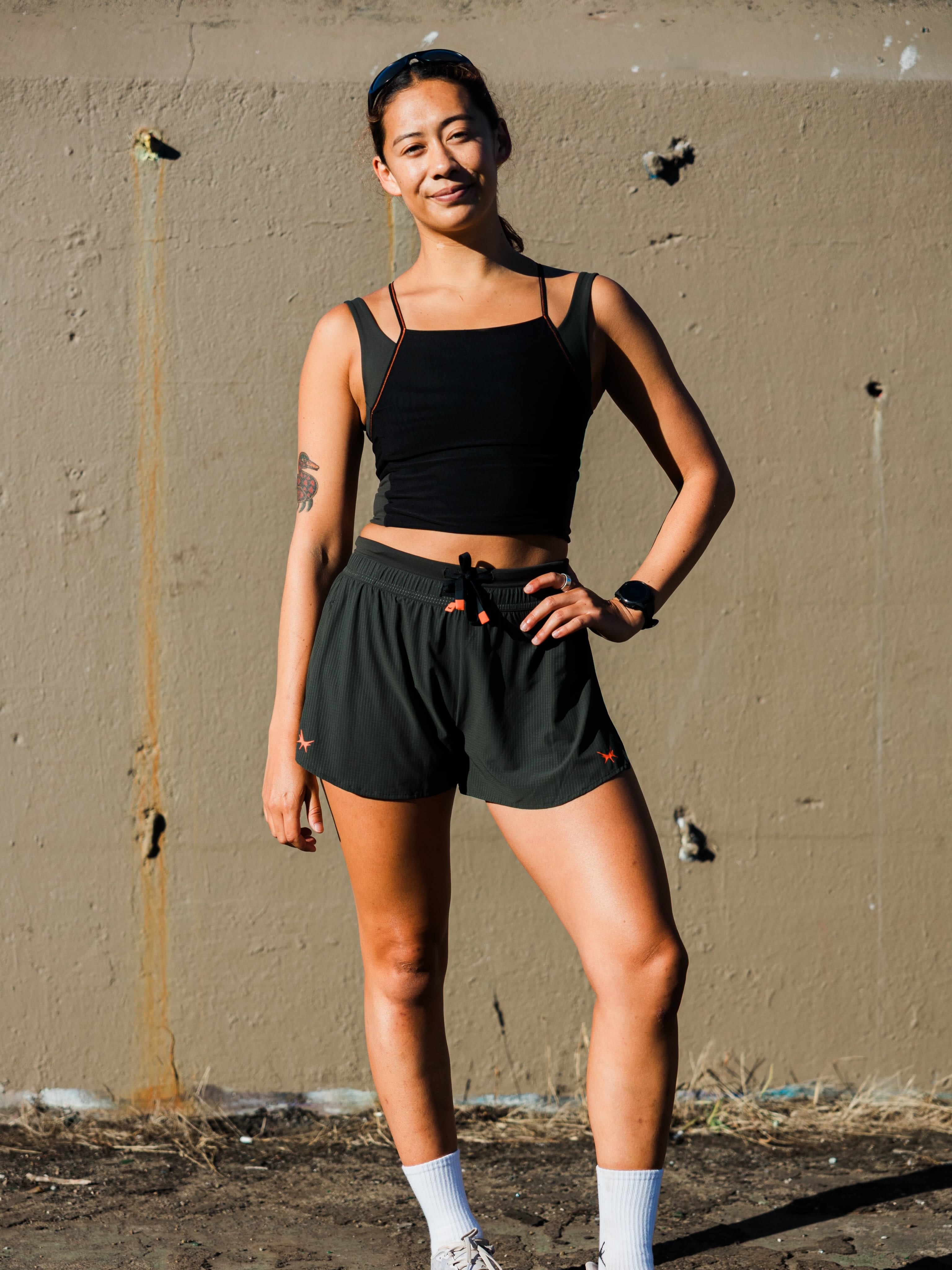 A woman in a black crop top and dark green athletic shorts stands against a textured concrete wall.