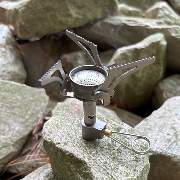 A small portable camping stove sits atop a grey rock surrounded by larger stones.