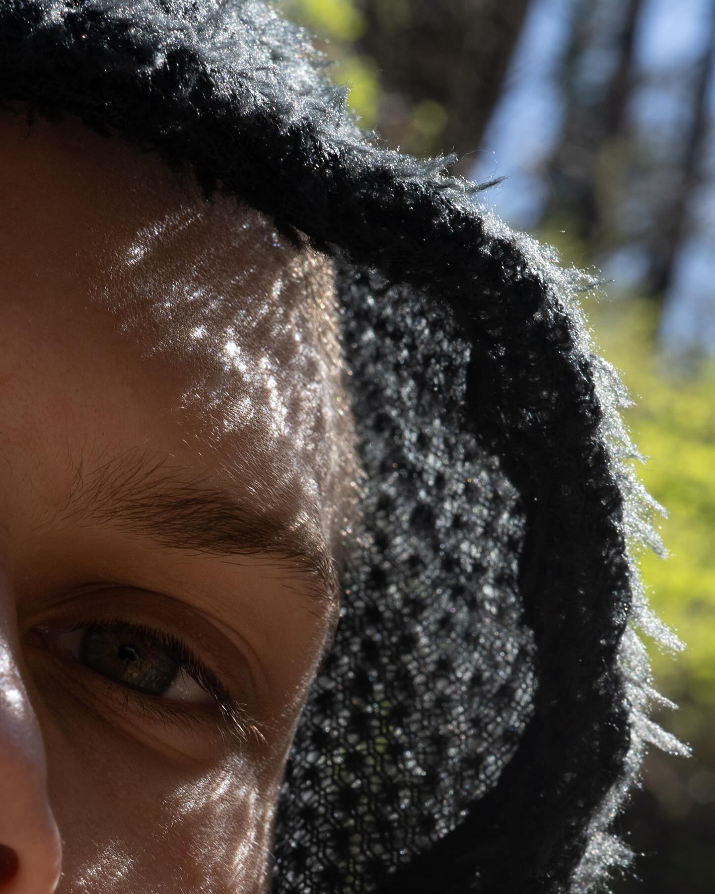 A close-up of a person's eye and forehead framed by a textured black hood, with a blurred background of green foliage and blue sky.