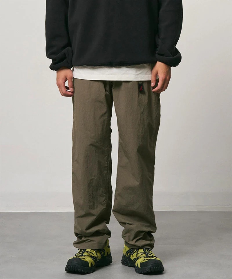 A person stands in loose-fitting olive pants and colorful hiking shoes against a plain gray background.