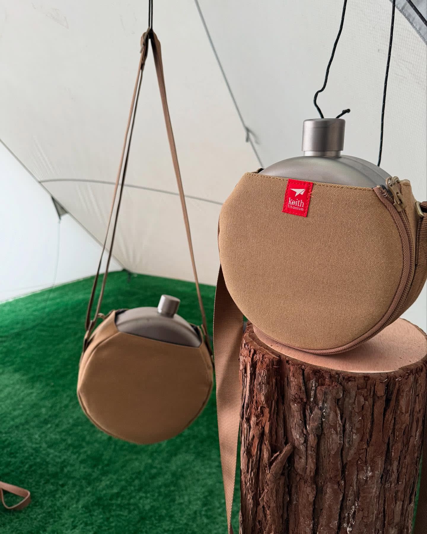Two tan kettle carriers, one resting on a wooden stump and another hanging, are set against a white tent and green artificial grass.