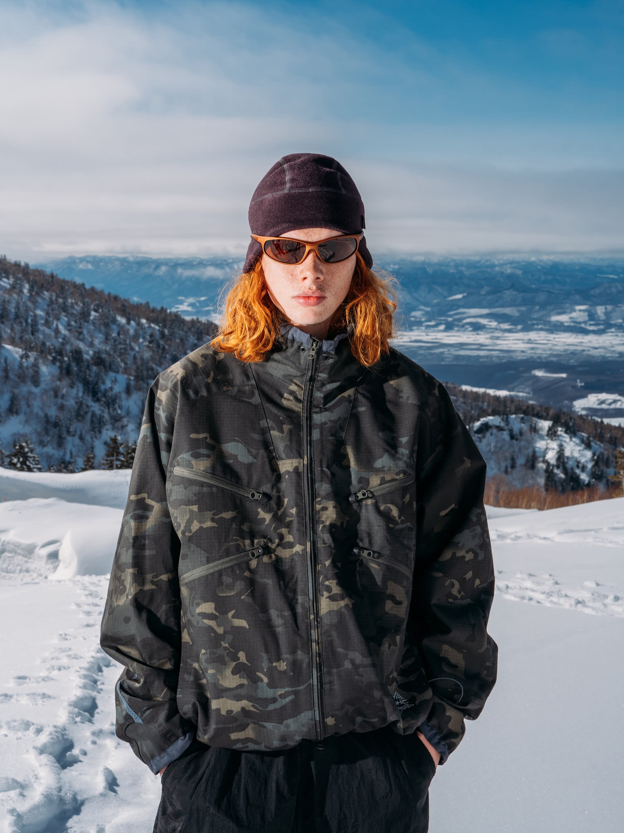 A person wearing a camouflage jacket, beanie, and sunglasses stands in the snowy foreground against a backdrop of distant mountains and a blue sky.