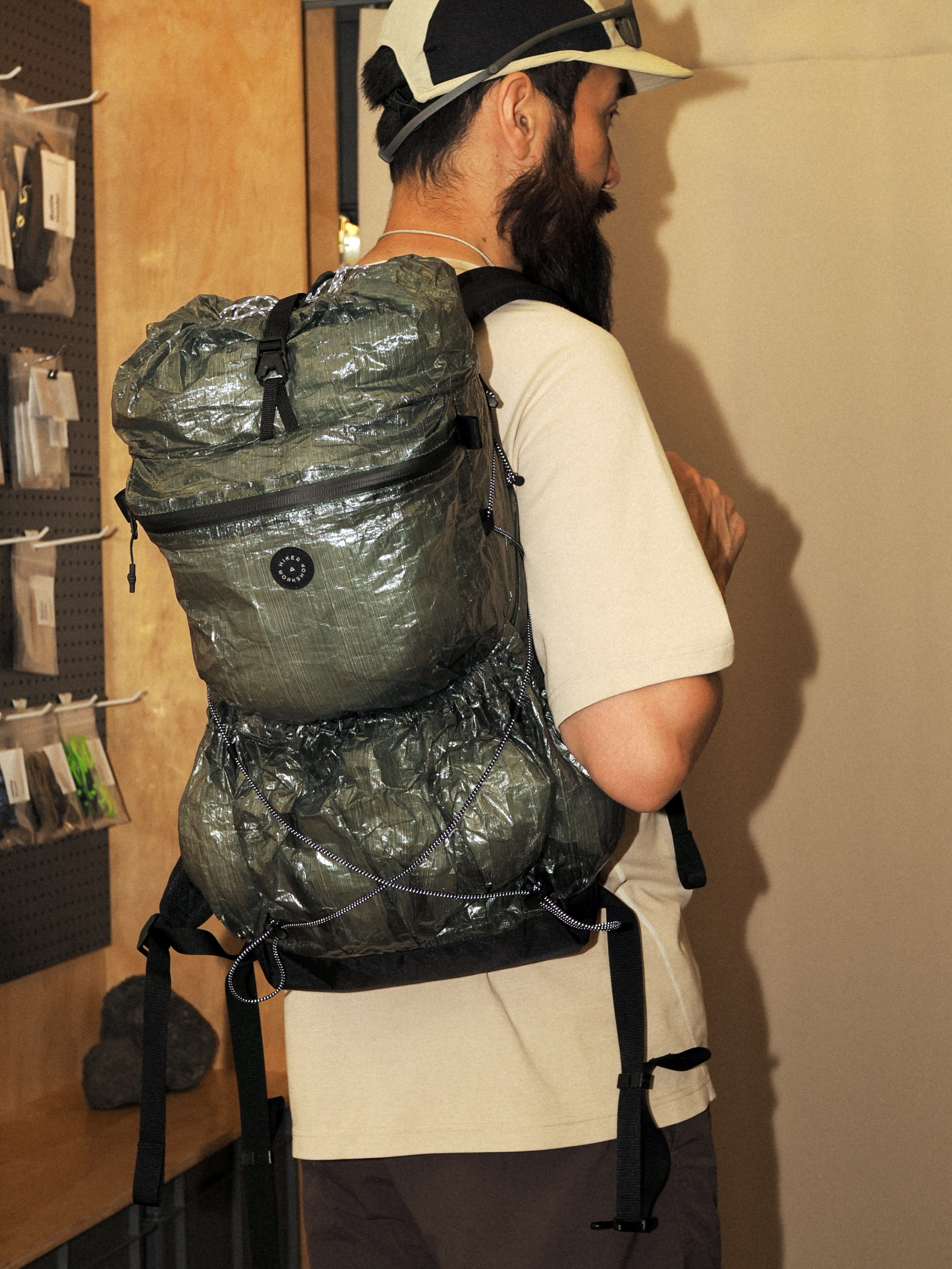 A man wearing a white t-shirt and a cap carries a large, crinkled grey backpack while standing in a room with a pegboard wall.