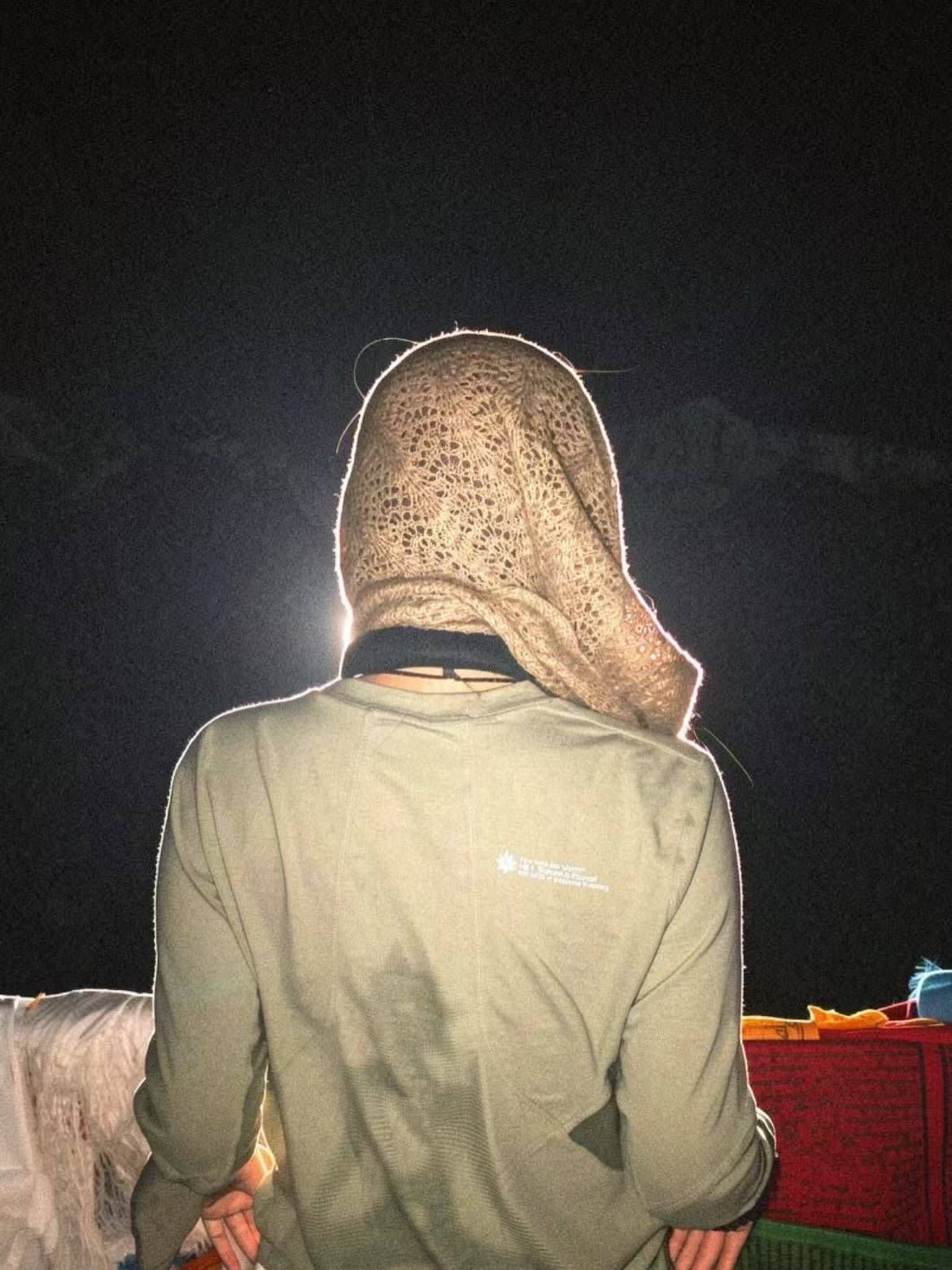 A person wearing a green shirt and a textured beige head covering stands against a dark, nighttime background.