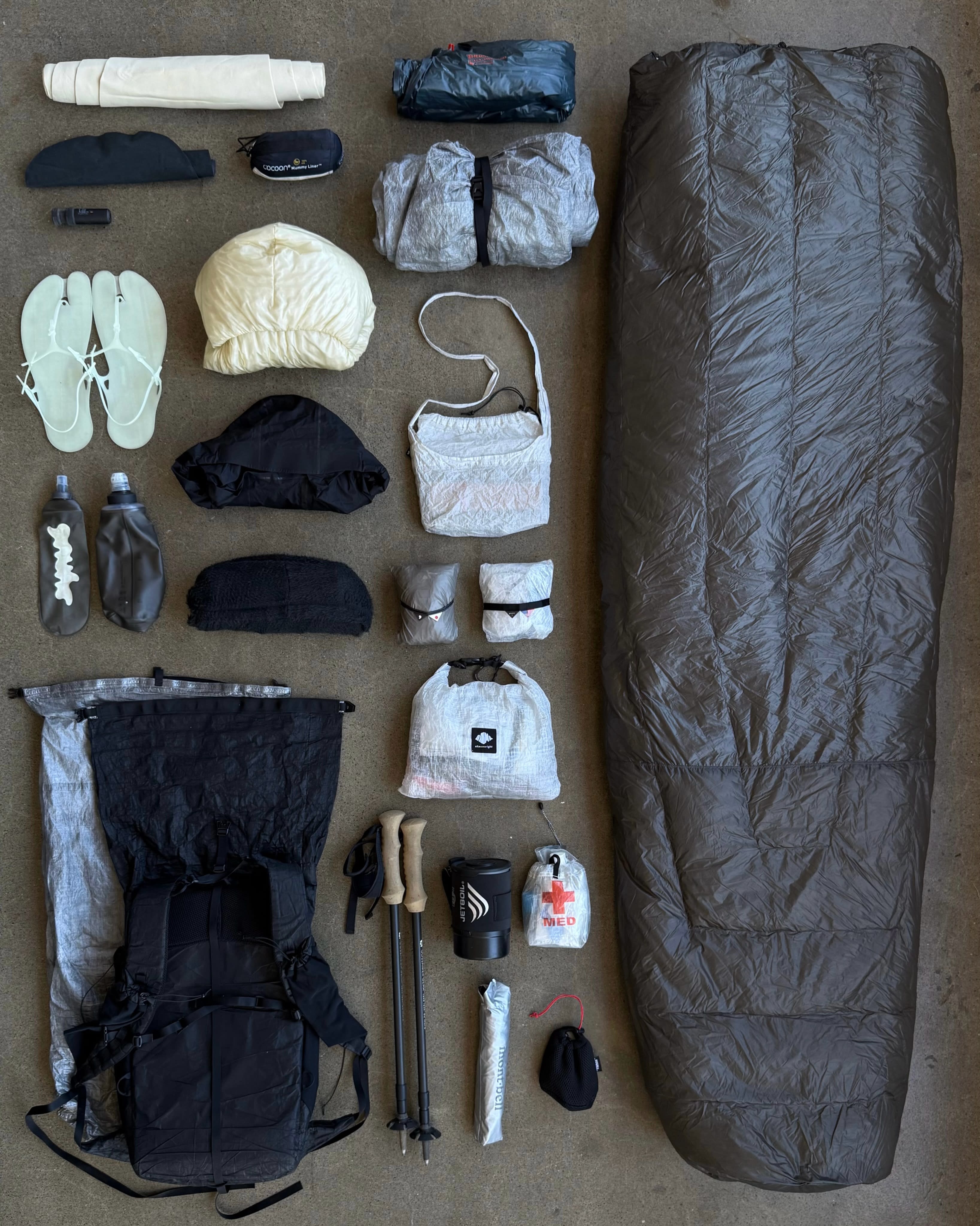 Various camping gear and hiking equipment, including a sleeping bag, backpack, and water bottles, are laid out on a flat gray surface.