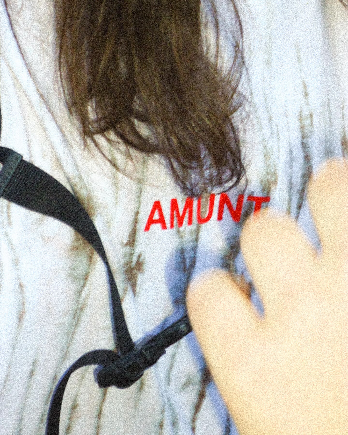A hand and a black strap are in the foreground against a white t-shirt featuring red text and a blurred background.