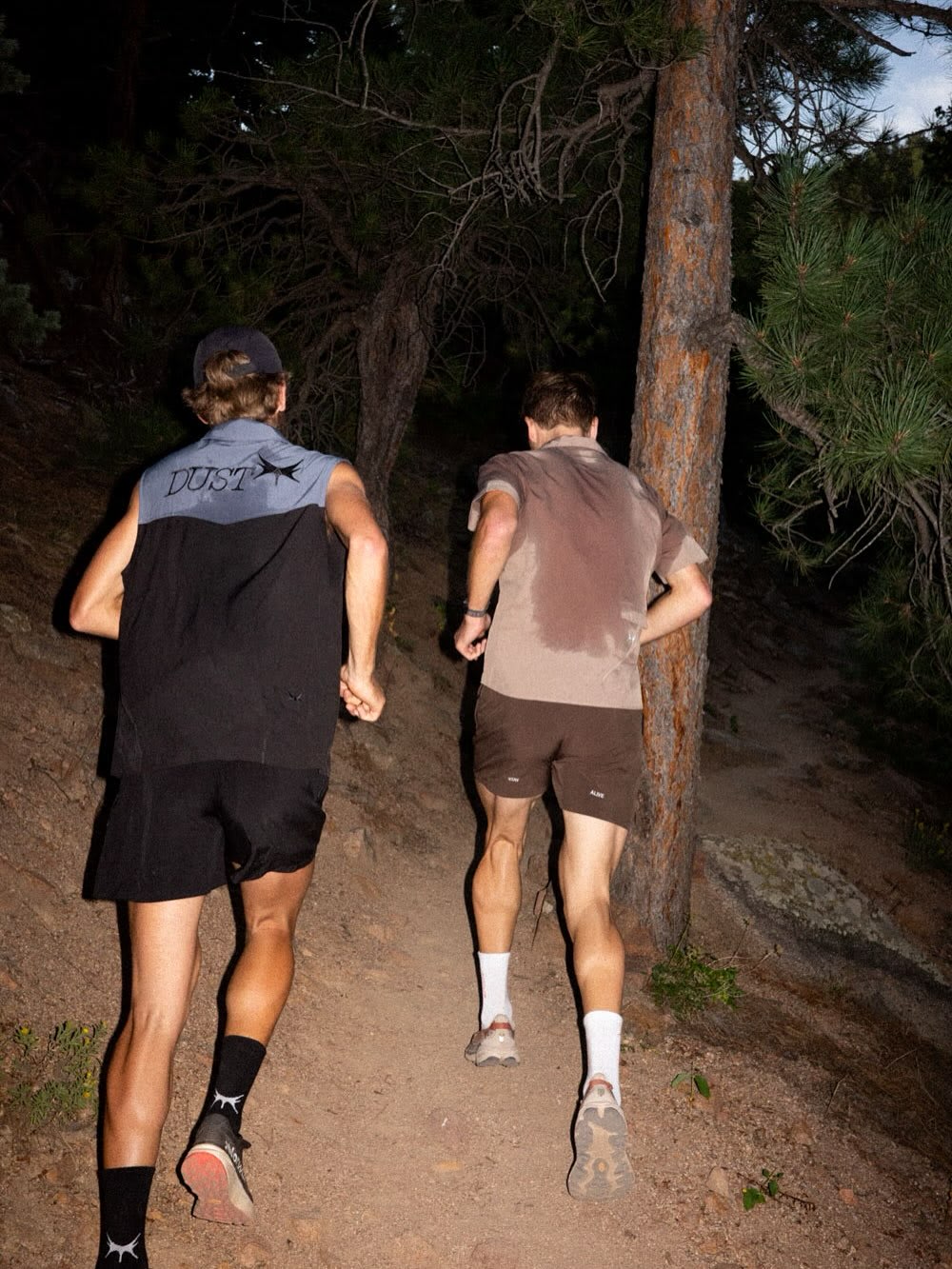 A blurred person's leg is in the foreground, while two people run through green brush under a clear blue sky in the background.