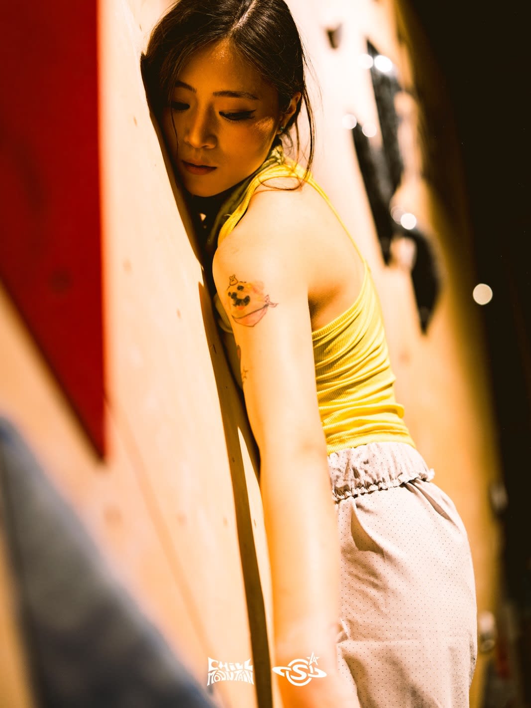 A woman in a yellow tank top leans against a light-colored climbing wall with dark holds in the background.