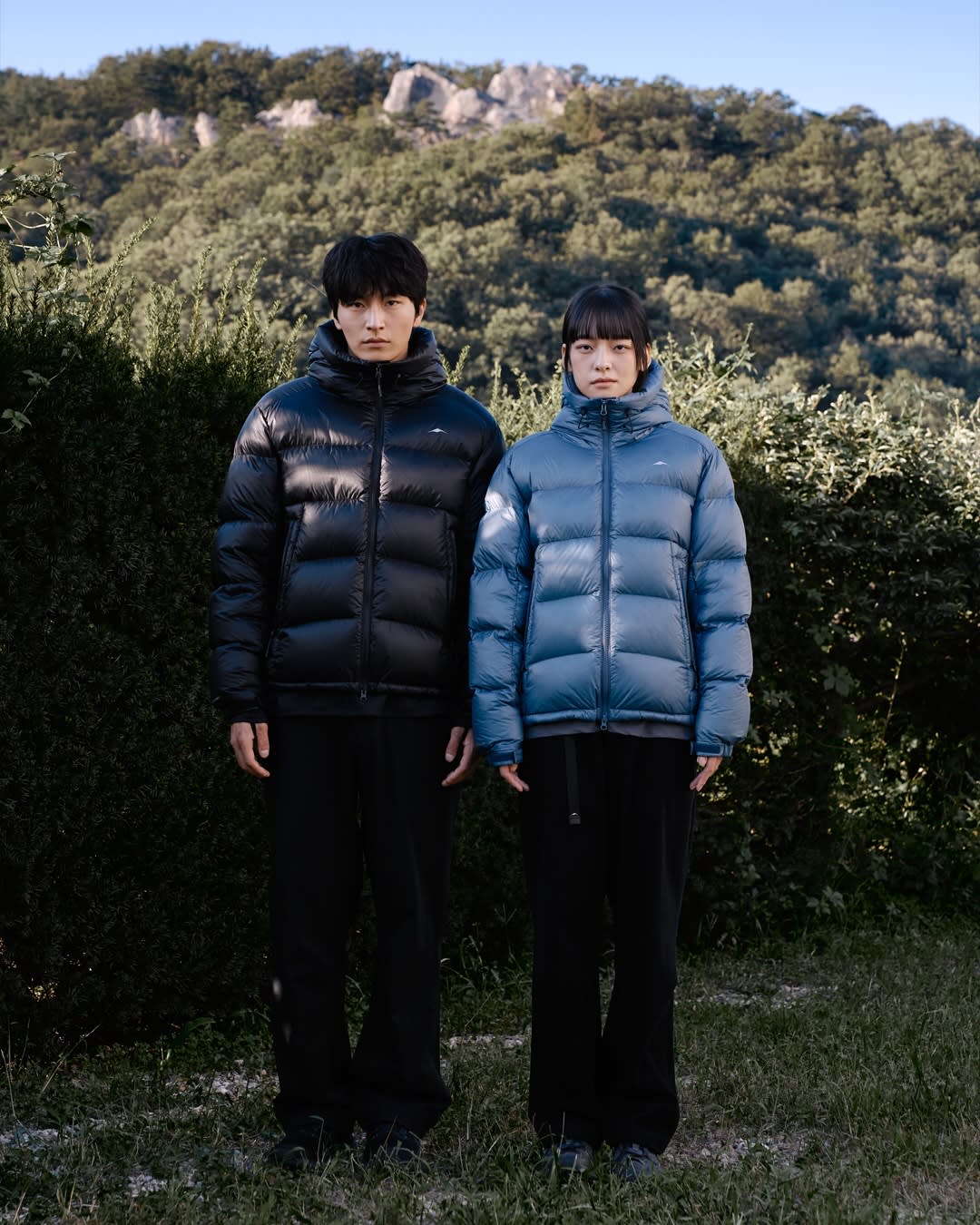 Two people wearing puffer jackets stand in a grassy area against a backdrop of dense green trees and rocky hills.