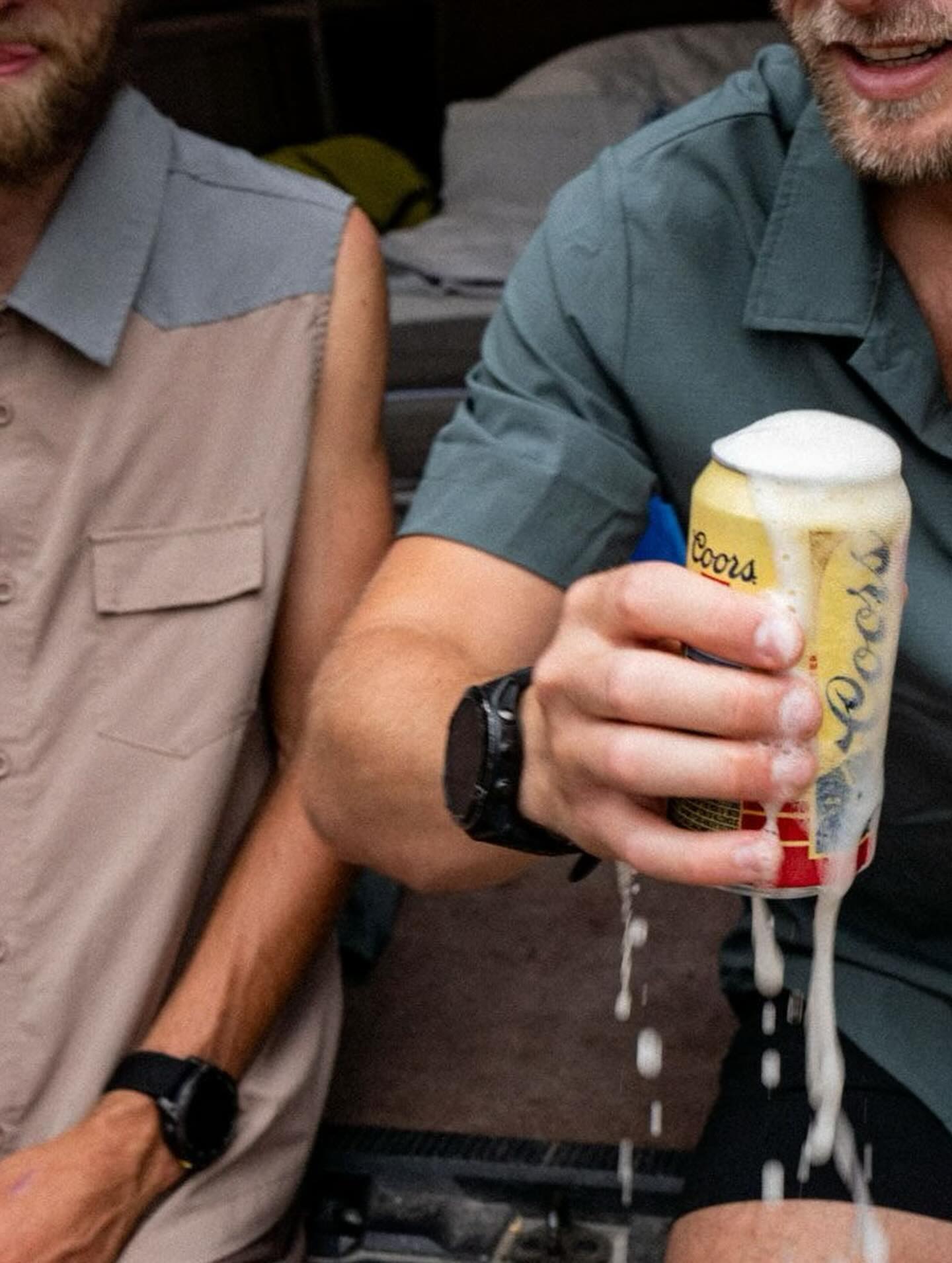 A man in a green shirt holds a foaming can of Coors beer in the foreground, while another man in a tan vest stands beside him against a blurred outdoor background.
