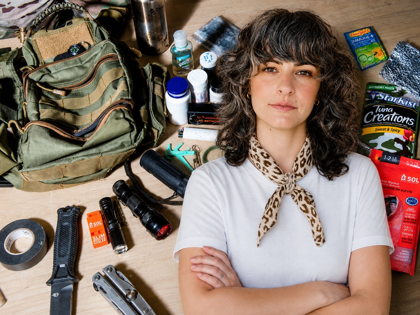 A woman with curly hair and a leopard-print scarf stands with her arms crossed, surrounded by a collection of survival gear including a backpack, flashlights, and canned food.