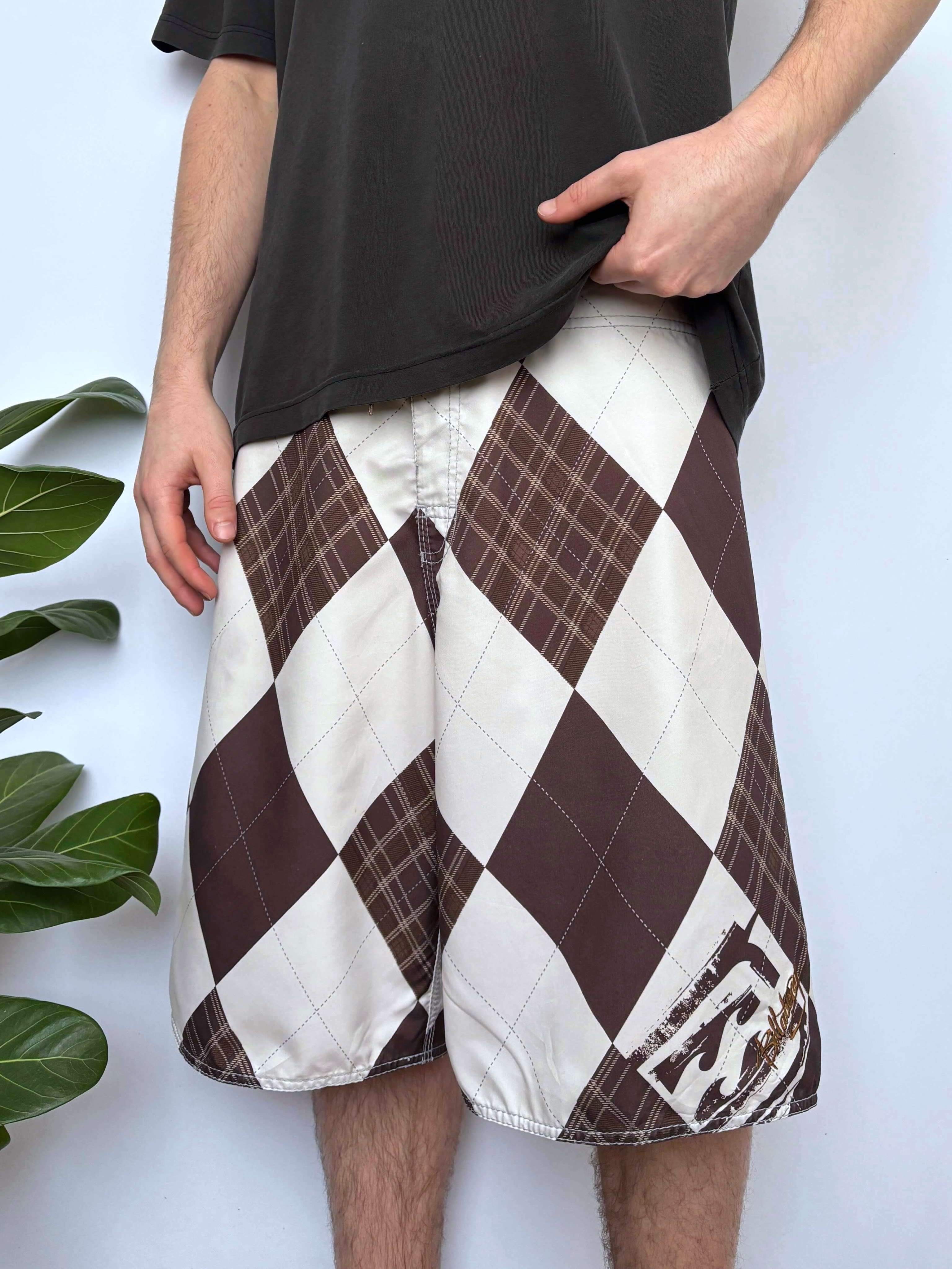 A person wearing a black t-shirt and brown and white diamond-patterned shorts stands against a plain white wall with a green plant to the left.
