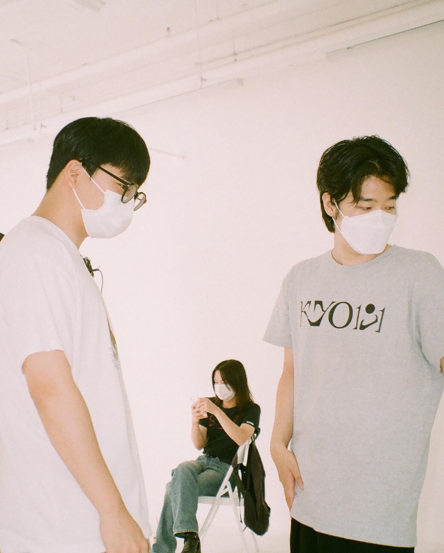 Three people wearing white face masks stand and sit in a minimalist white room.