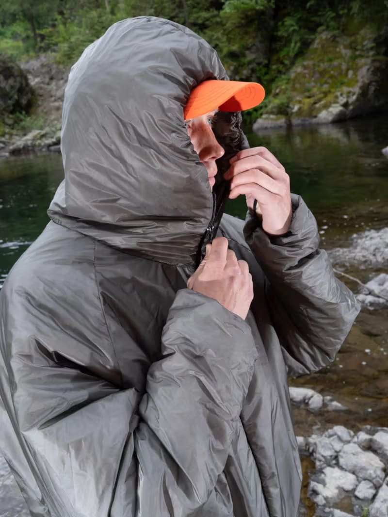 A person wearing a grey hooded puffer jacket and an orange cap stands before a river and rocky shoreline.