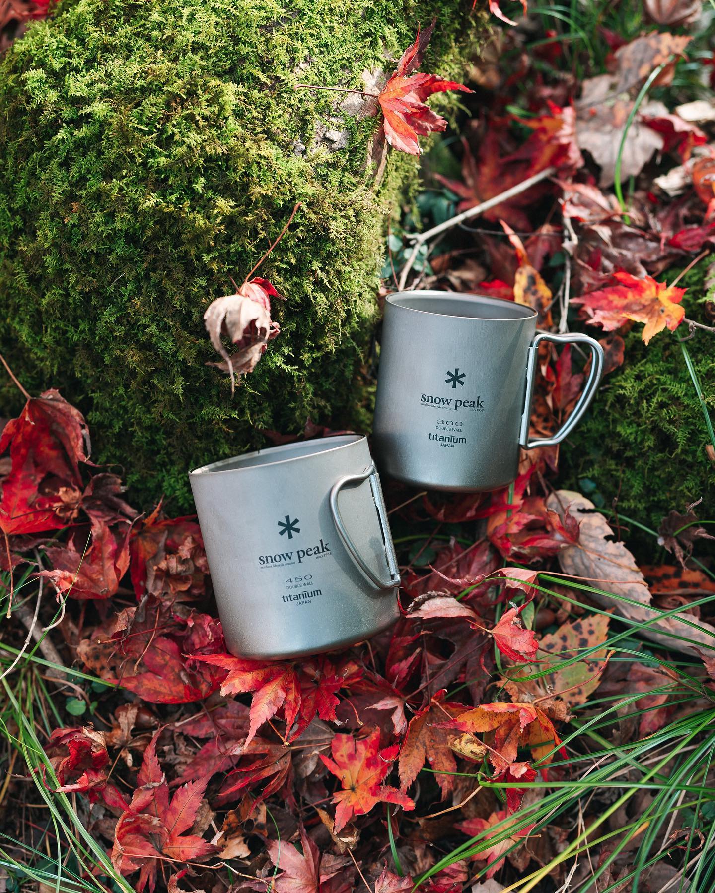 Two silver Snow Peak titanium mugs sit atop a bed of red autumn leaves in front of a moss-covered rock.