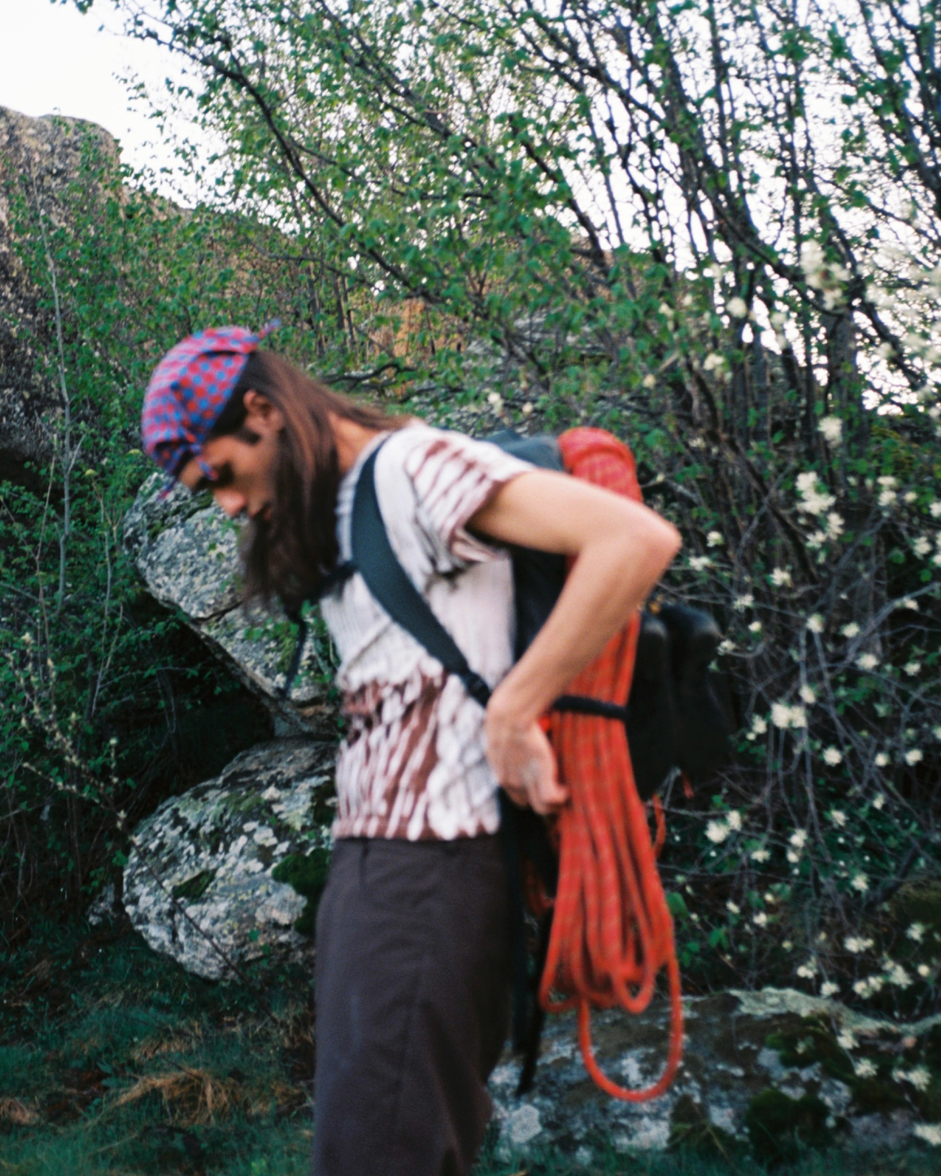 A person wearing a red patterned bandana and carrying a coil of orange climbing rope on their back stands before a backdrop of green foliage and rocky terrain.