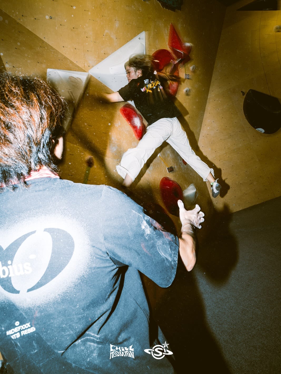 A man in a grey t-shirt watches from the foreground as another person climbs a yellow bouldering wall with red and white holds in the background.