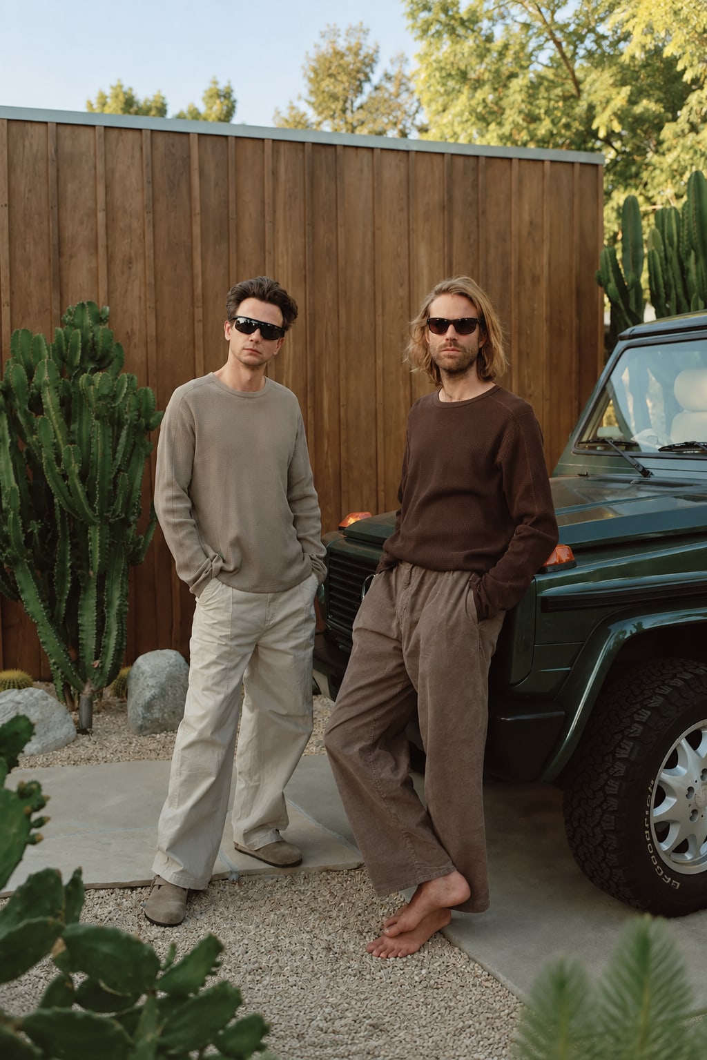 Two men wearing sunglasses and neutral-toned clothing stand beside a dark green off-road vehicle in front of a wooden fence and desert greenery.