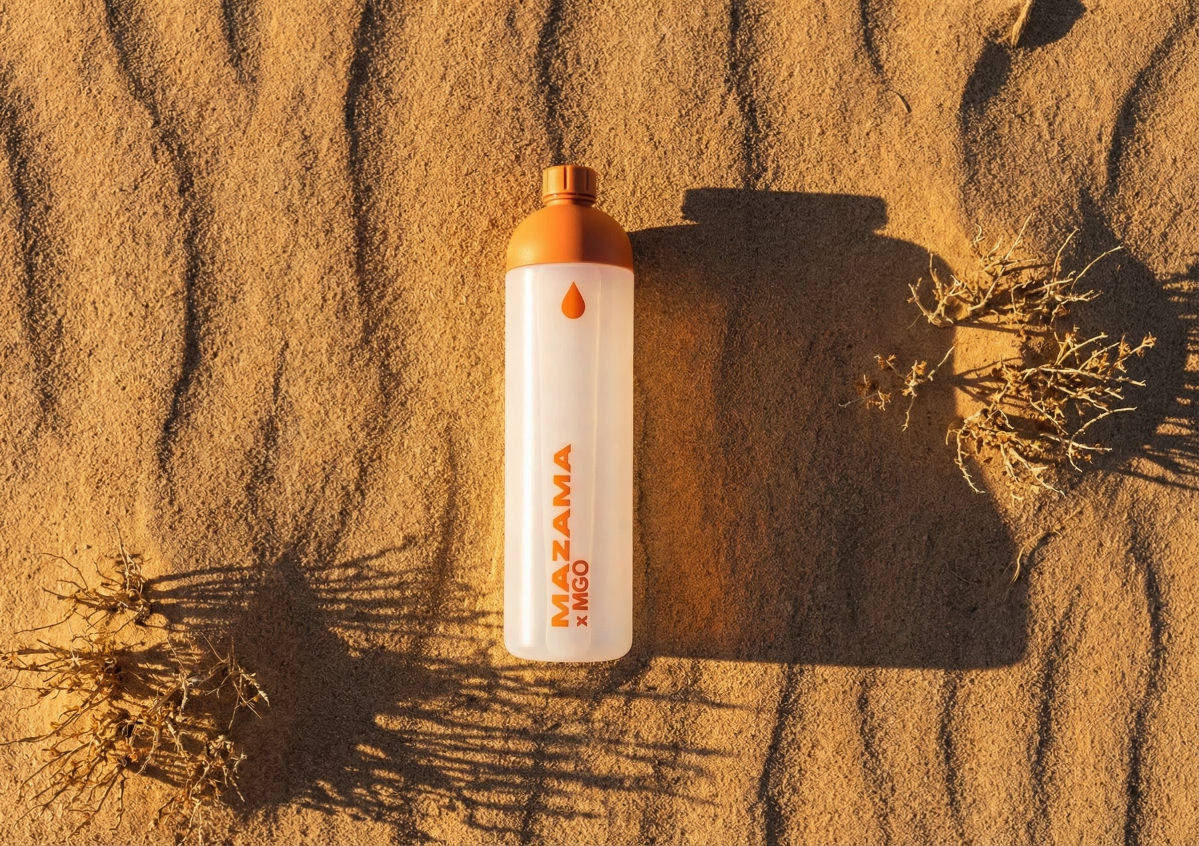 An orange and white water bottle stands upright on rippled desert sand, flanked by small, dried shrubs.